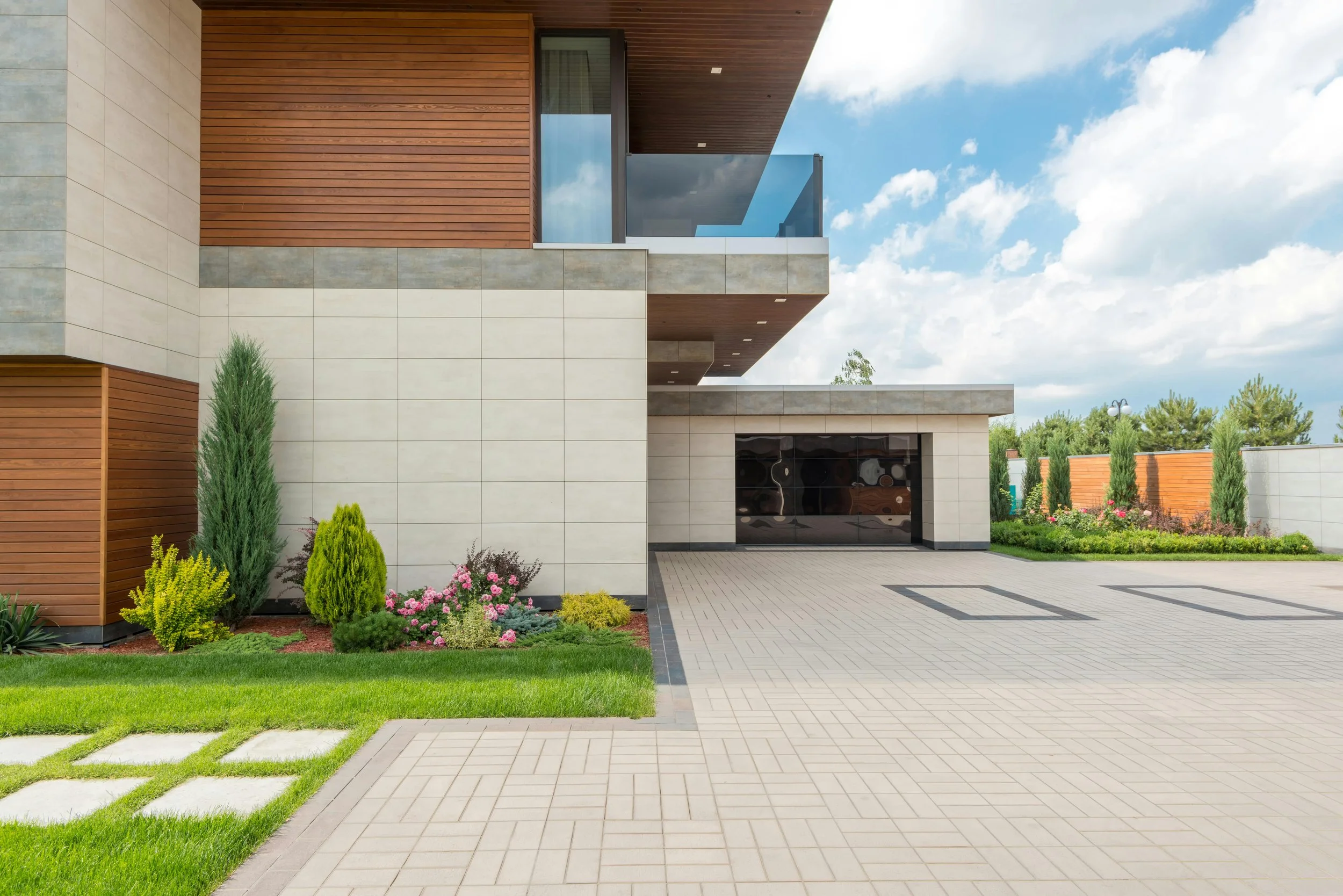 Modern house with a mix of beige stone, brown wood siding, and glass balcony, with landscaped front yard featuring green grass, bushes, and flowers, under a partly cloudy sky.