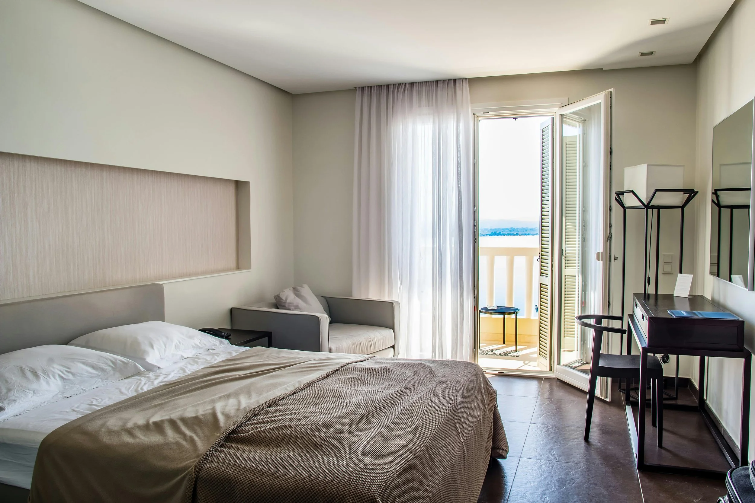 A hotel room with a large bed, a small sofa, a balcony door with sheer curtains, and a view of a body of water in the distance. There is a small black desk and a mirror on the wall.