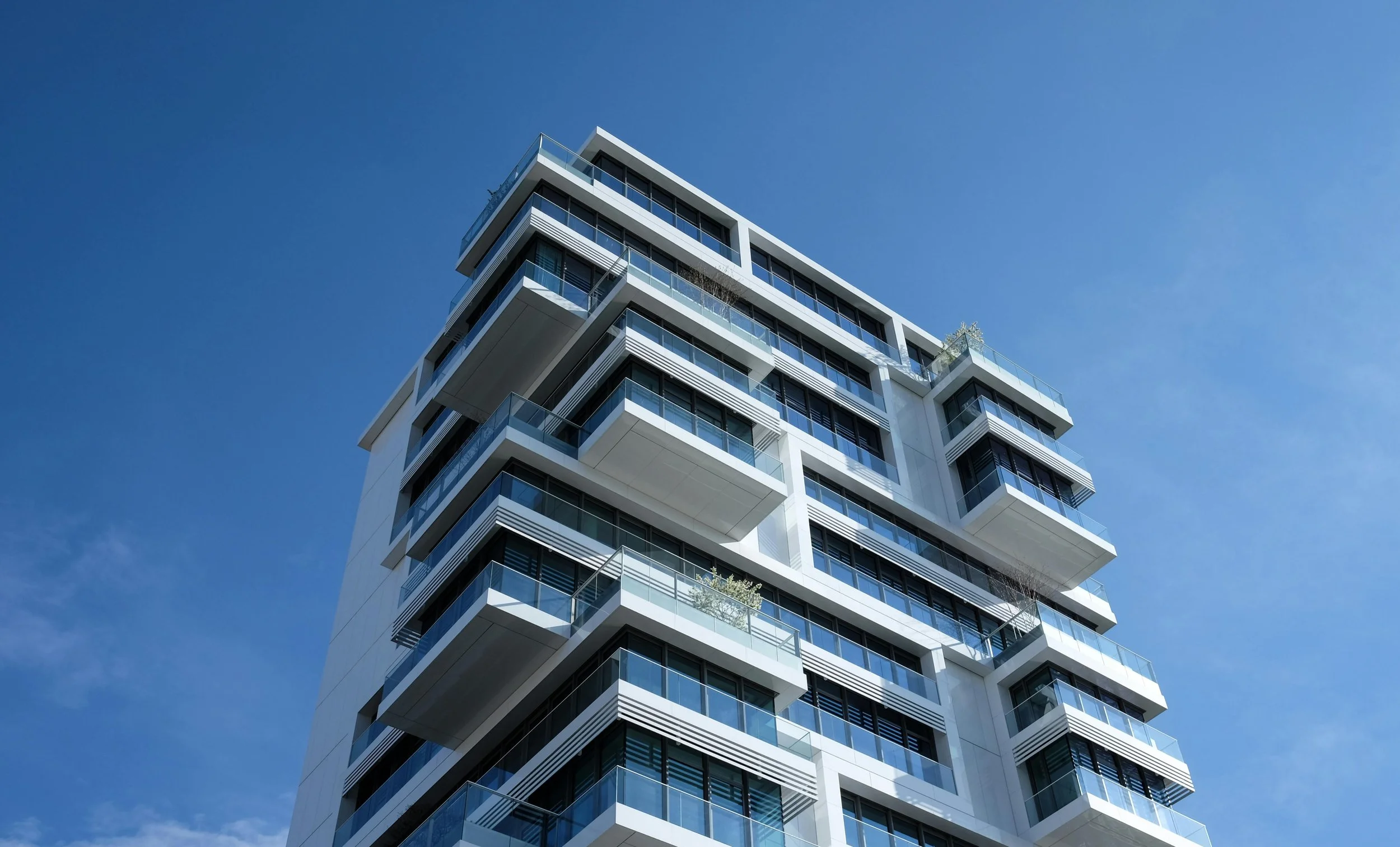 A modern high-rise building with multiple balconies and glass railings, set against a clear blue sky.