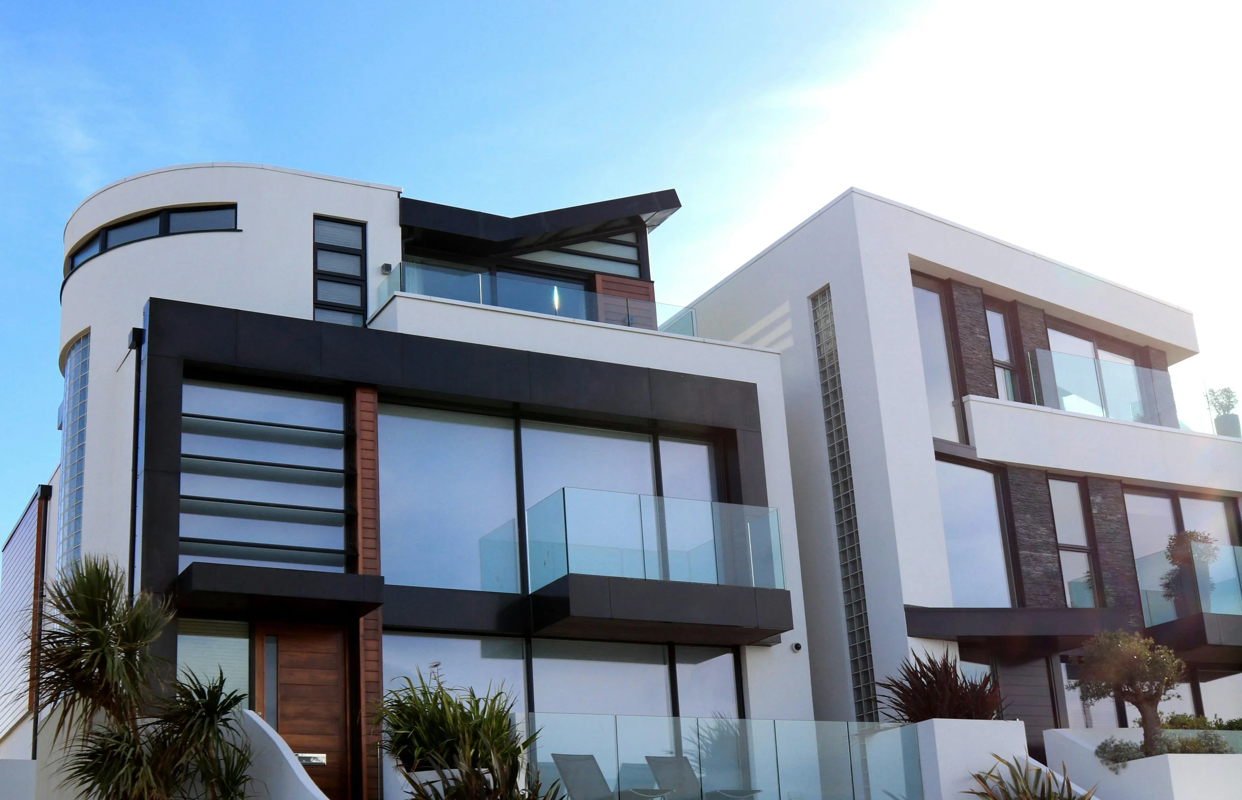 Modern multi-story building with large glass windows and balconies, with landscaping including trees and plants in the foreground, under a bright blue sky.