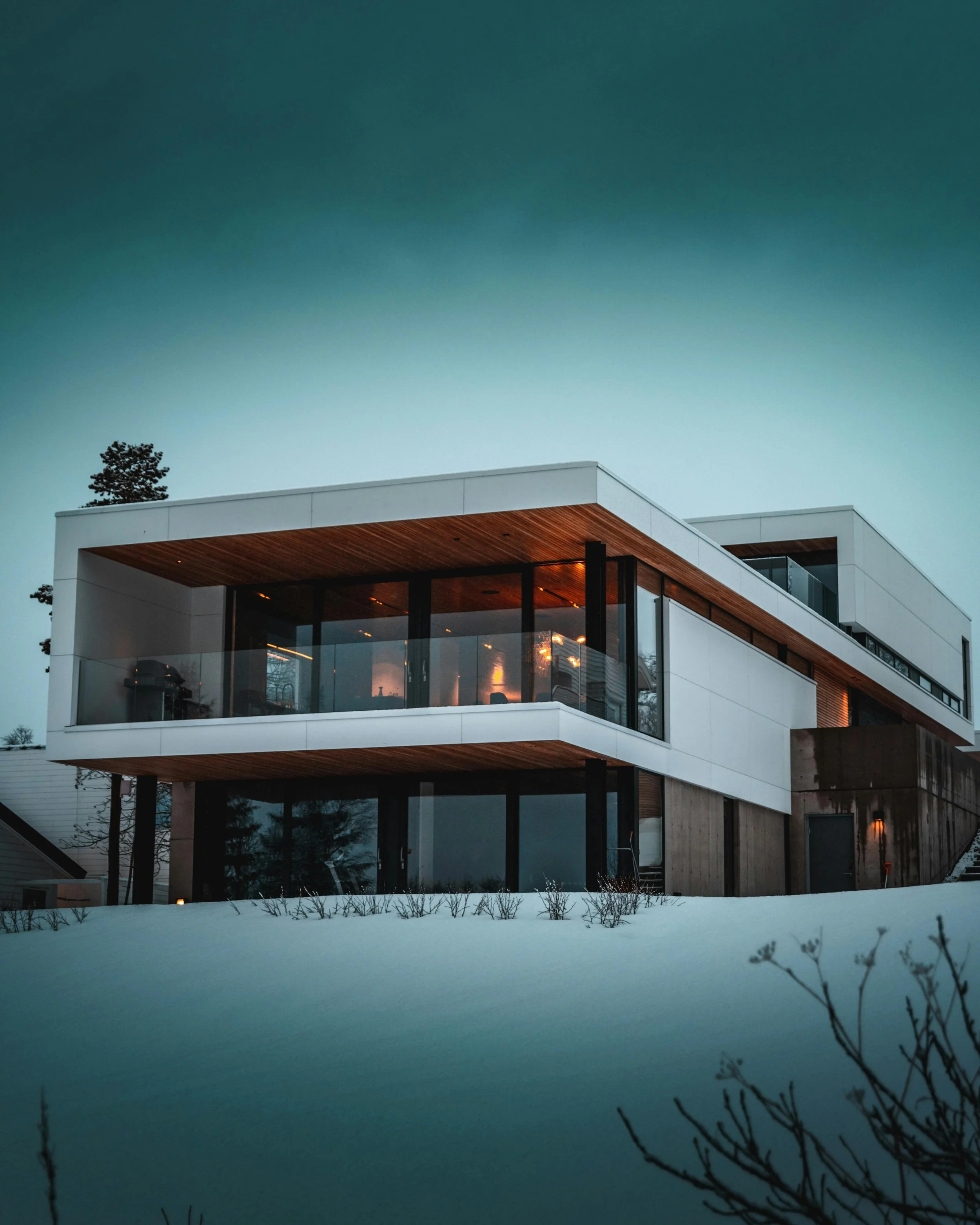Modern multi-story house with large windows and wooden accents, illuminated from inside, set against evening sky with snow on the ground and silhouettes of trees and bushes in the foreground.