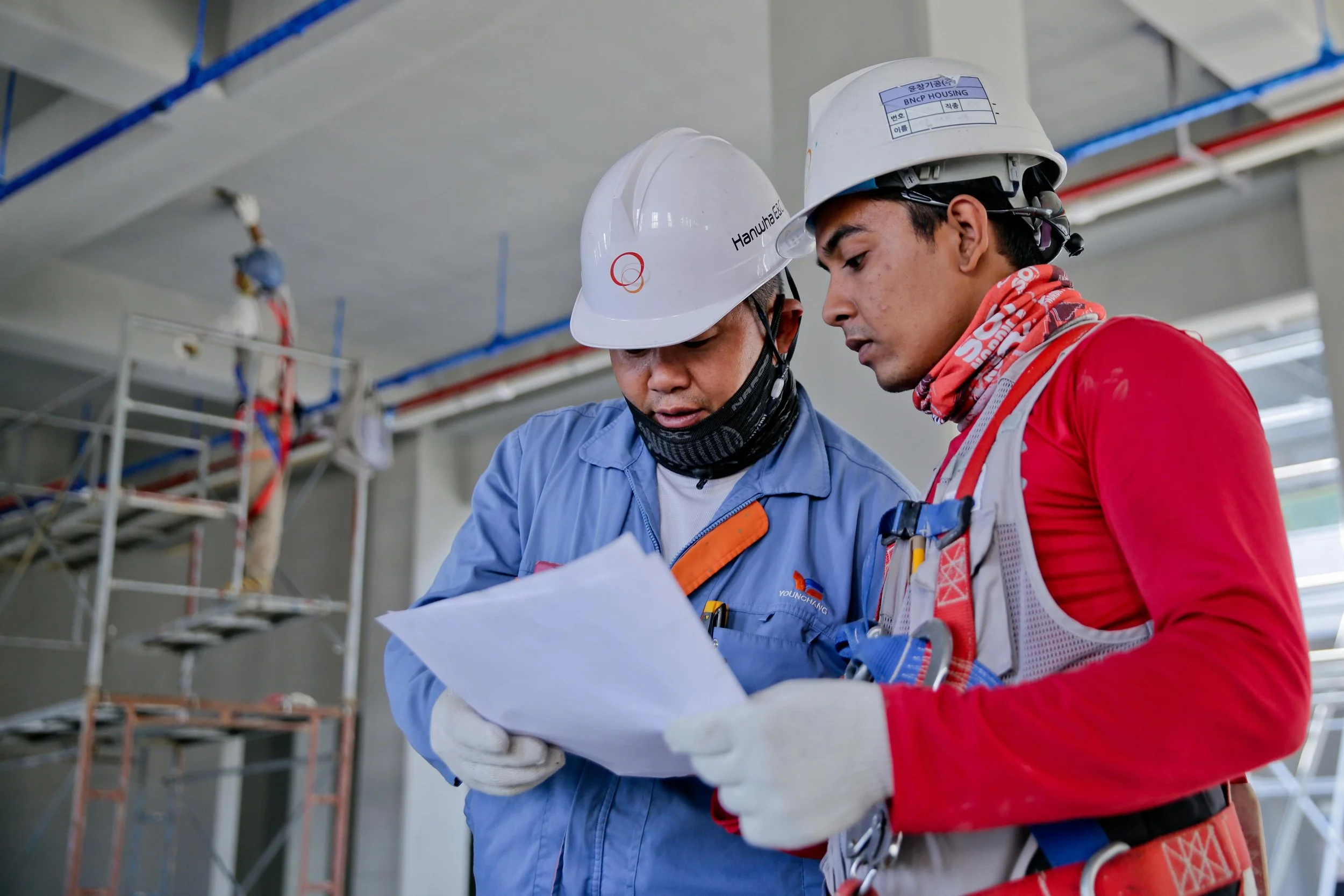 Two construction workers wearing helmets and safety gear reviewing documents on a construction site.