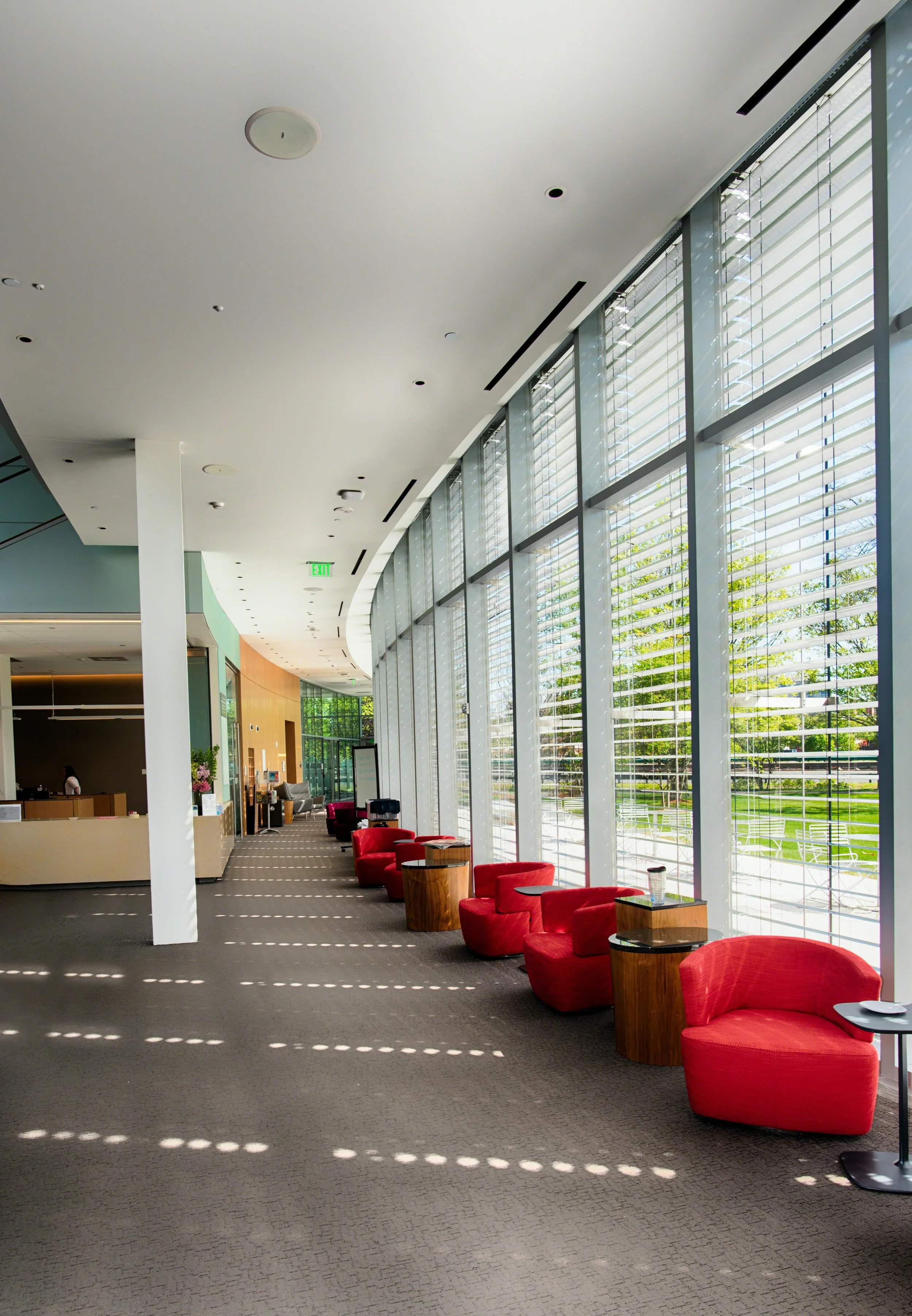 Modern interior of a bright, spacious lounge or waiting area with large floor-to-ceiling windows and red armchairs lined along the window. Outside the window, greenery and trees are visible. There is a reception desk and a person sitting behind it in the background.