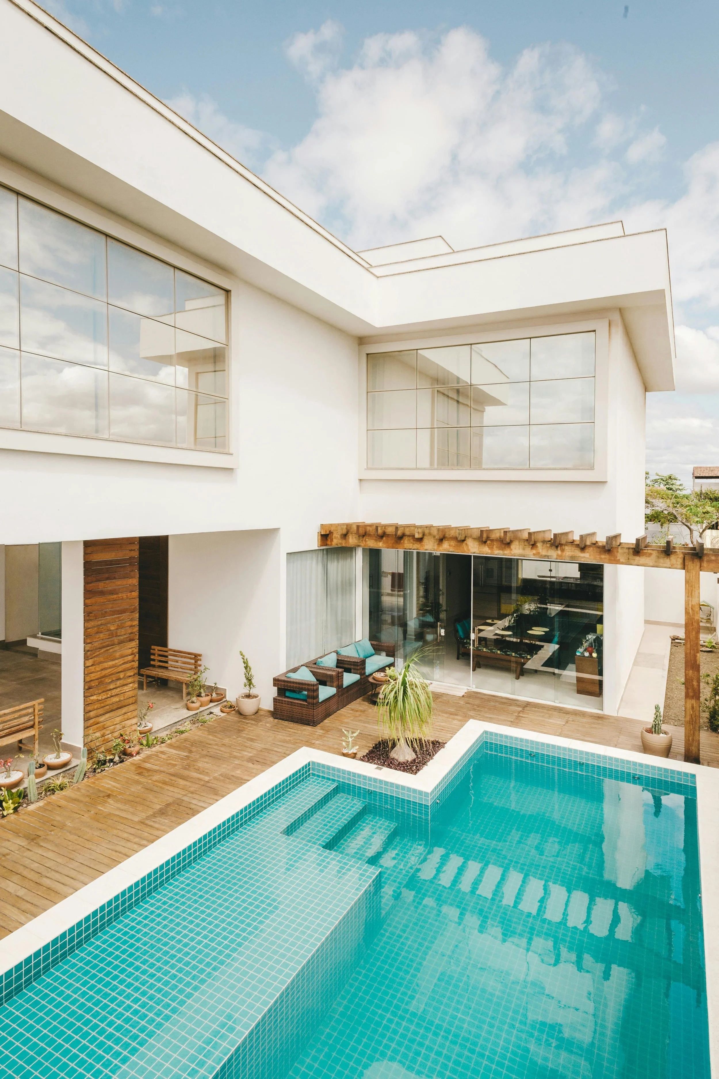Modern house with large glass windows, a small swimming pool with light blue tiles, potted plants, outdoor seating, and wooden decking, under a partly cloudy sky.