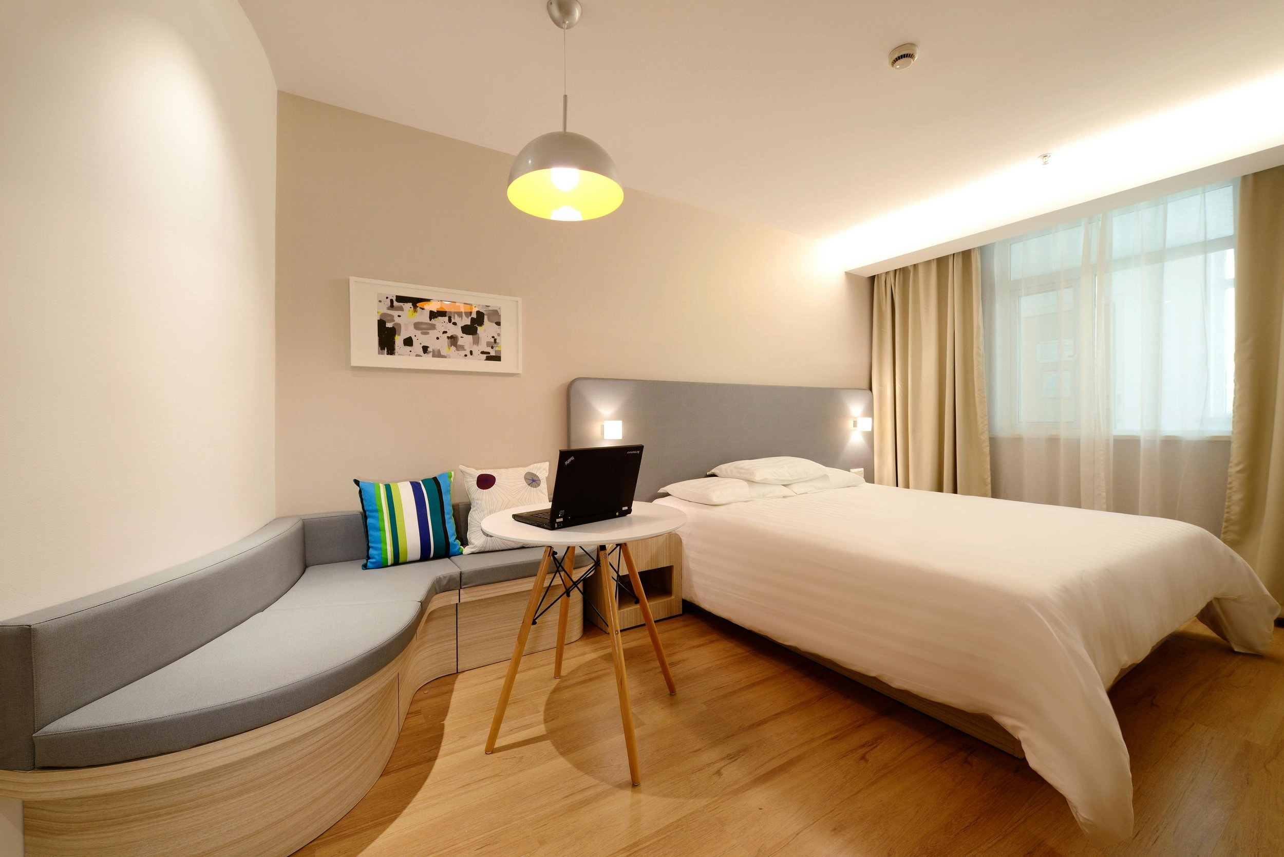 A hotel room with a large bed, a circular desk with a laptop, colorful pillows, a gray curved seating area, abstract artwork on the wall, a hanging ceiling light, and a large window with curtains.