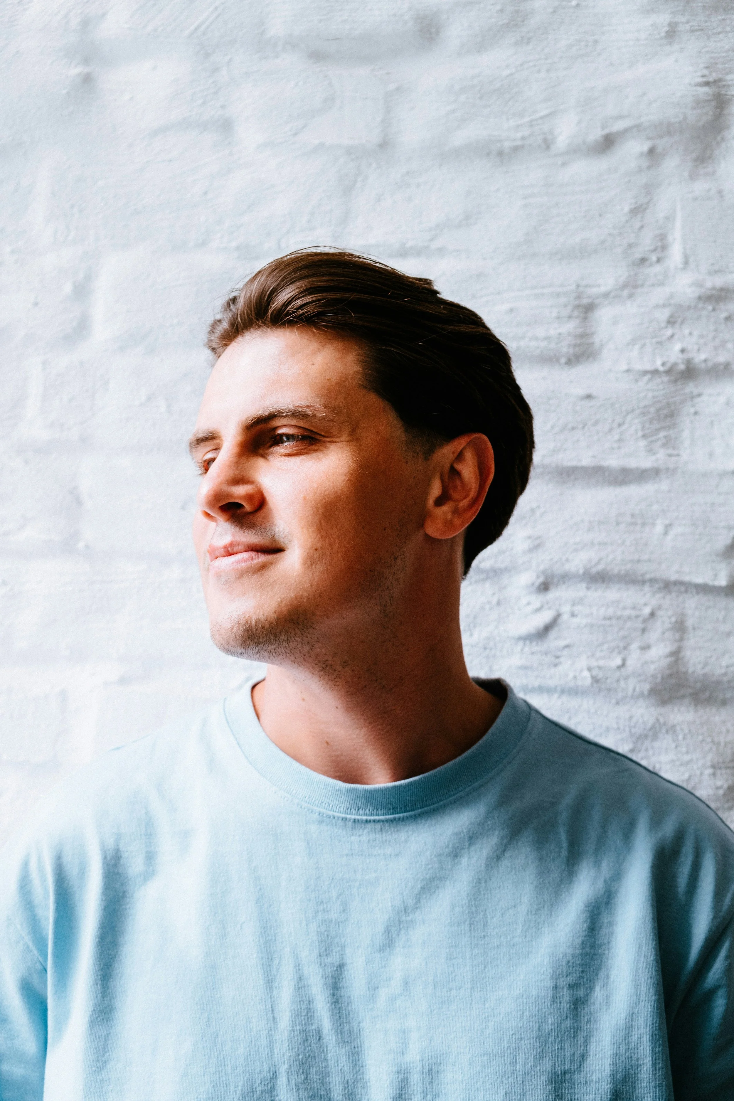 Close-up of a young man with brown hair, wearing a light blue T-shirt, standing against a white brick wall, looking slightly to the side with a confident expression.