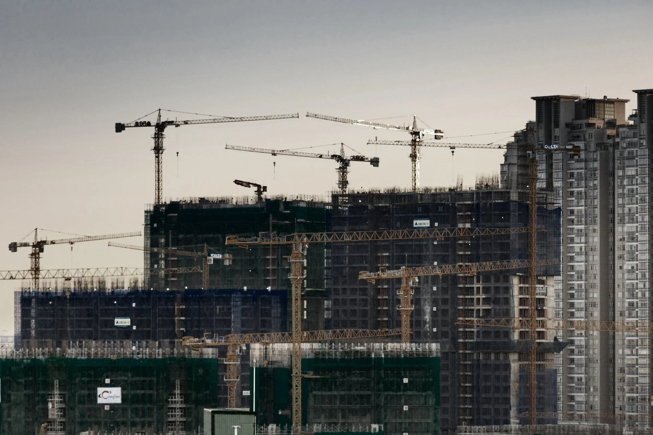 A construction site with multiple cranes and a high-rise building under construction, with some completed high-rise buildings in the background under a gray sky.