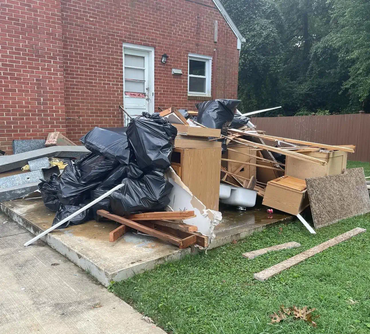 Debris and trash piled outside a brick house, including black garbage bags, wooden furniture, and construction materials.