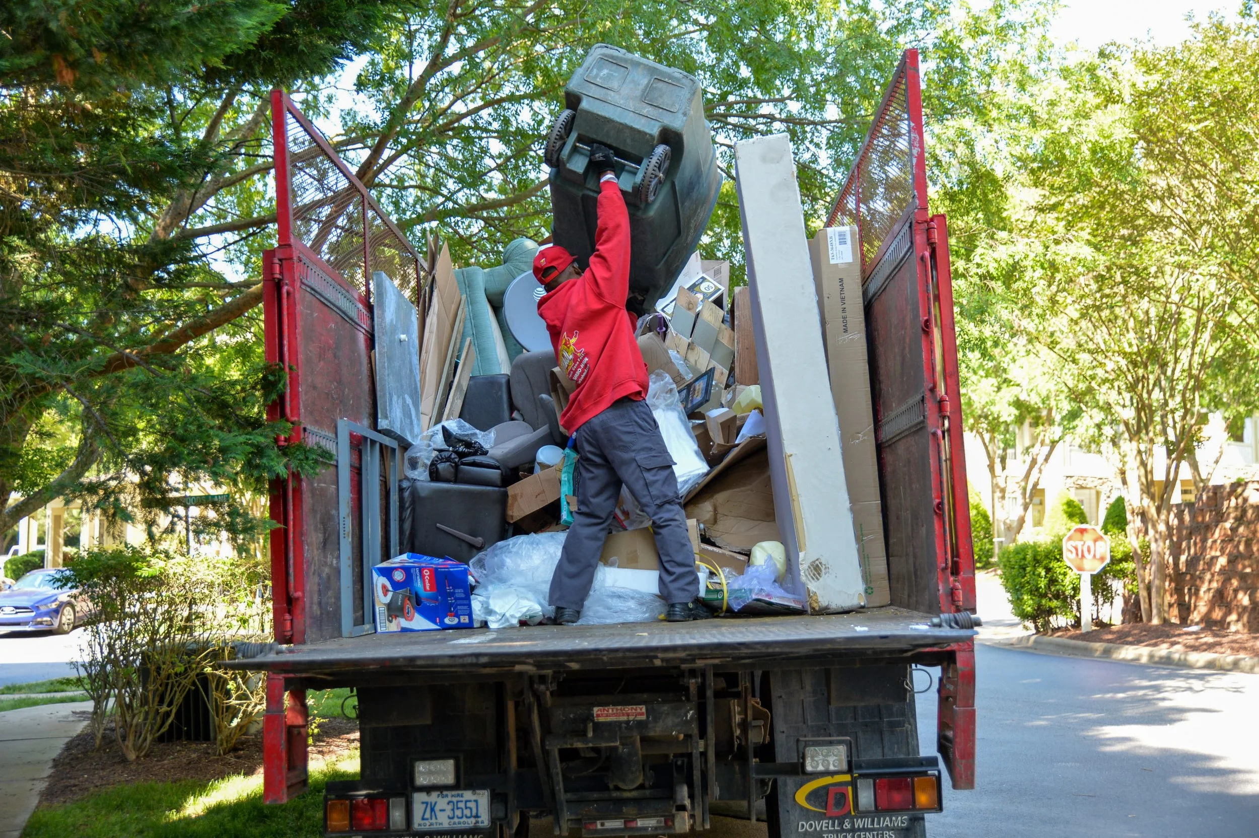 A person wearing a red jacket and a red cap loading trash and discarded items onto a red truck parked on a residential street surrounded by green trees
