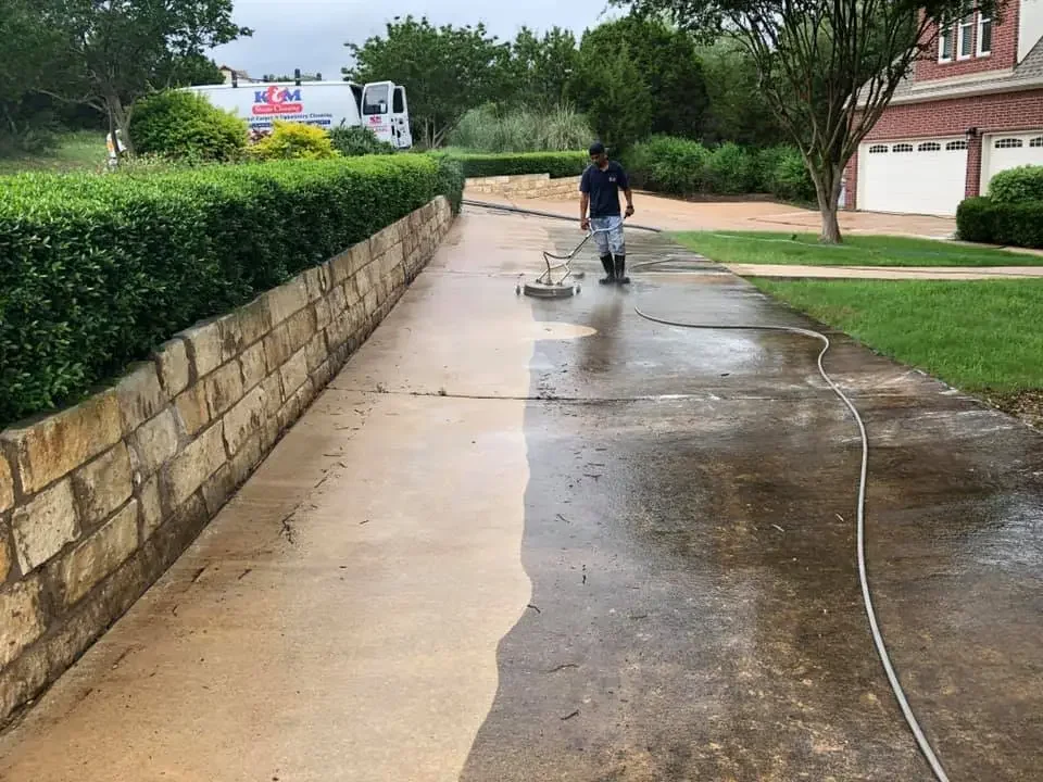 A person cleaning a driveway with a pressure washer, with the left side wet and clean and the right side still dirty, in a residential neighborhood with trees, shrubs, and houses.
