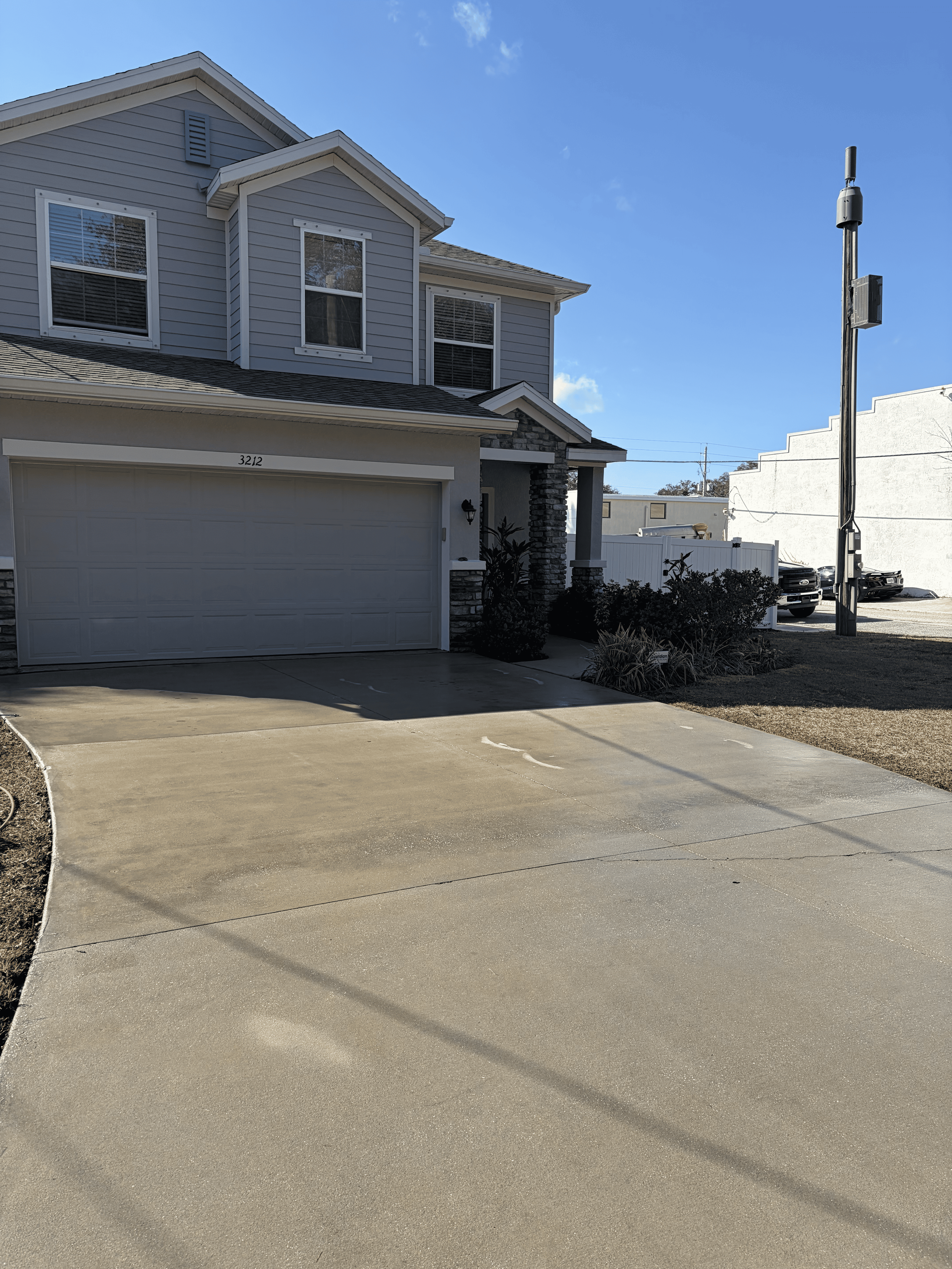 A two-story house with gray siding and a front garage door, with a clear blue sky overhead, shadows cast on the concrete driveway.