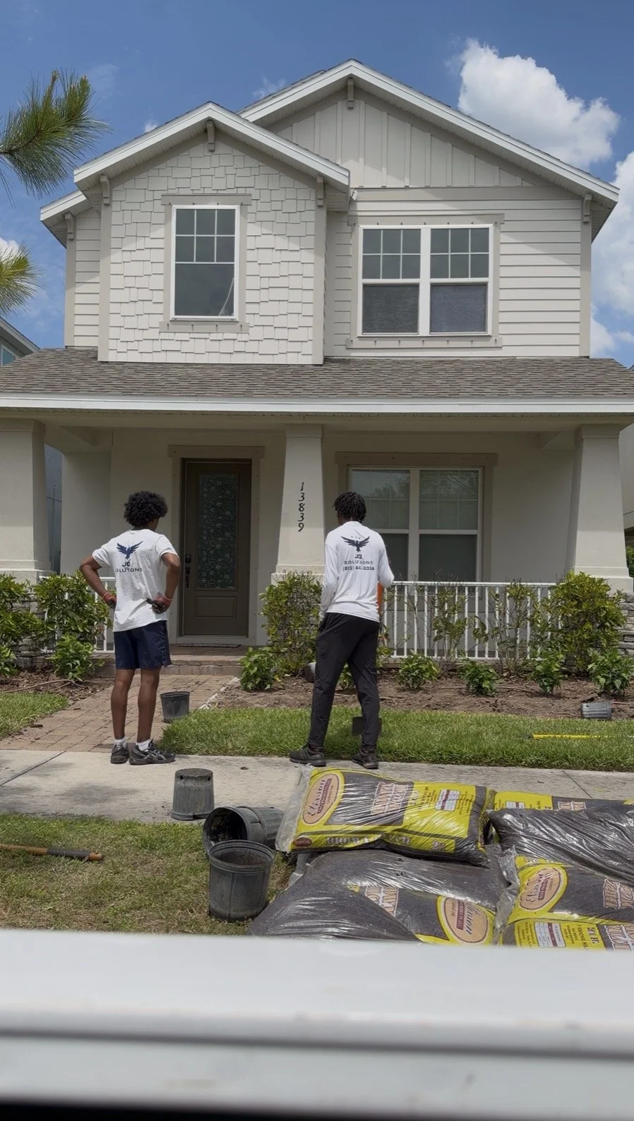 Two workers in uniform working on landscaping in front of a two-story house with a manicured lawn, shrubs, and a brick pathway, under a partly cloudy sky.