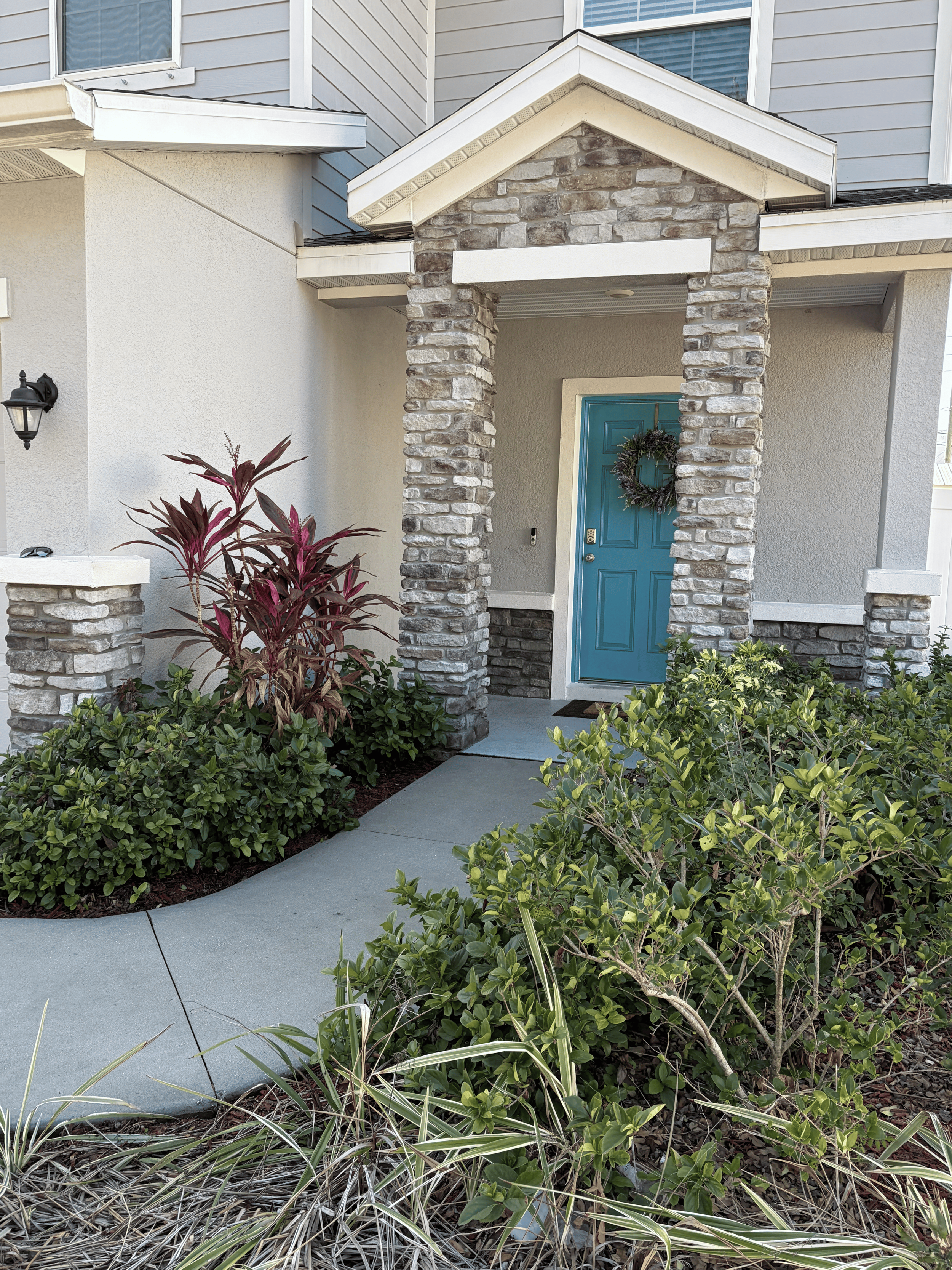 View of a house's front entrance with a blue door decorated with a Christmas wreath, surrounded by stone columns and greenery, with a concrete walkway leading to the door.