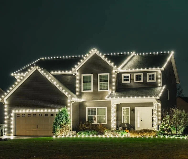 A house decorated with white Christmas lights outlining the roof, windows, and garage at night.