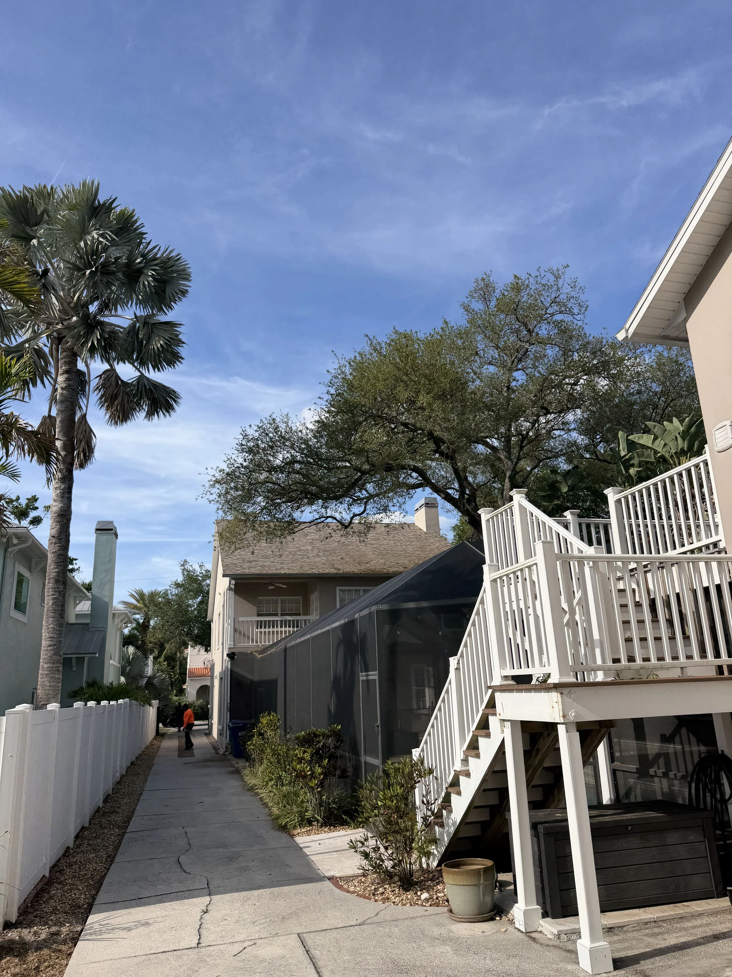 Residential backyard scene featuring a sidewalk, white fences, a two-story house, and tall trees with a clear blue sky.
