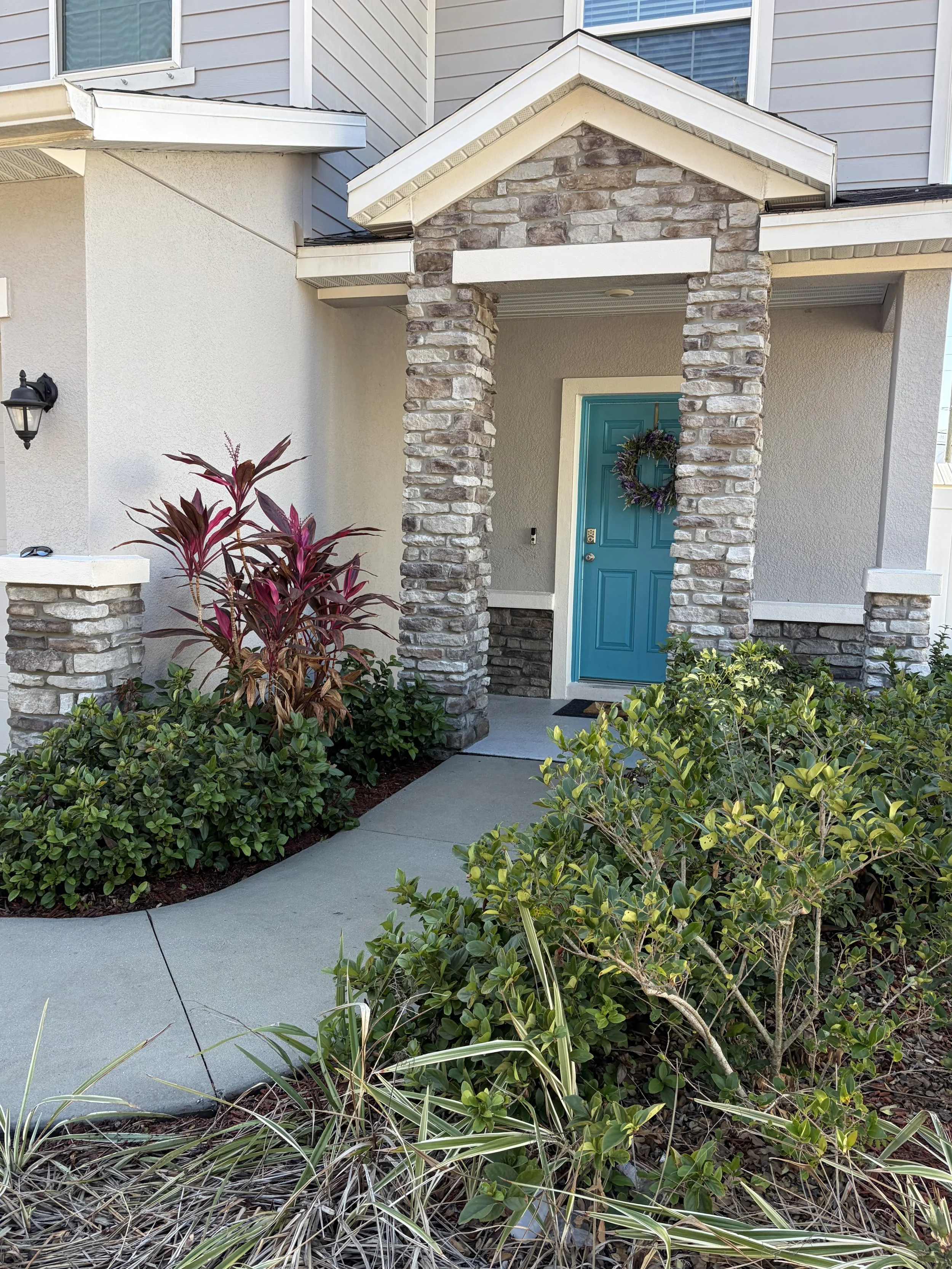 Front entry of a house with a blue door, brick and siding exterior, bushes and plants along the walkway, and a wreath on the door.