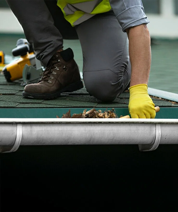 Close-up of a person caulking a roof, kneeling on shingles wearing work boots, yellow gloves, and work clothes, with roofing tools nearby.