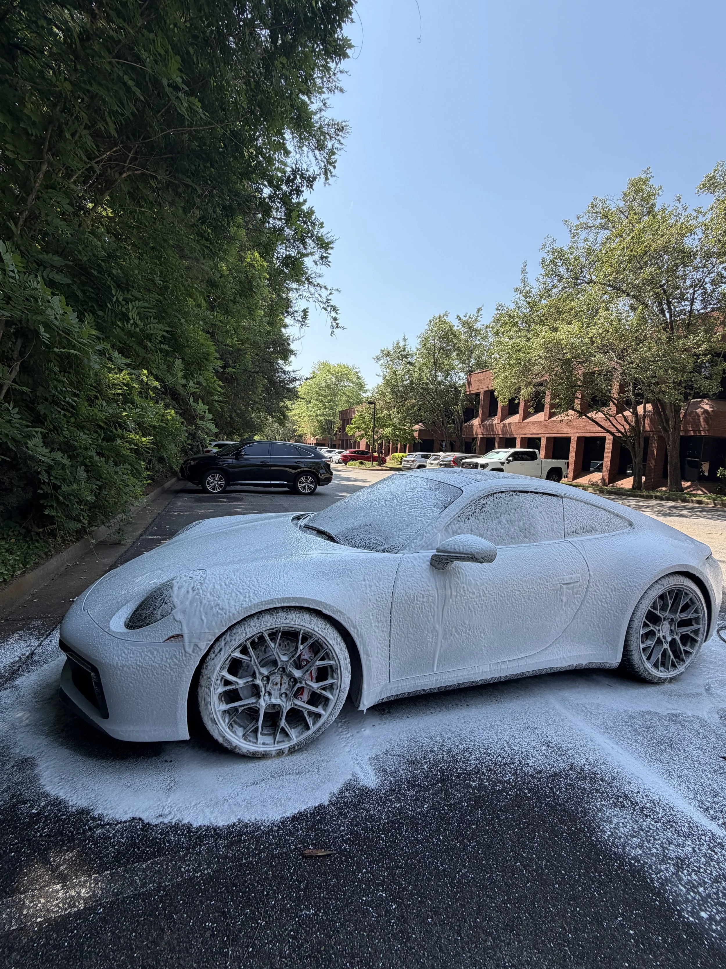 A white sports car covered in foam and soap, parked in a parking lot during daytime. The background includes other cars, a brick building, and green trees under a clear blue sky.