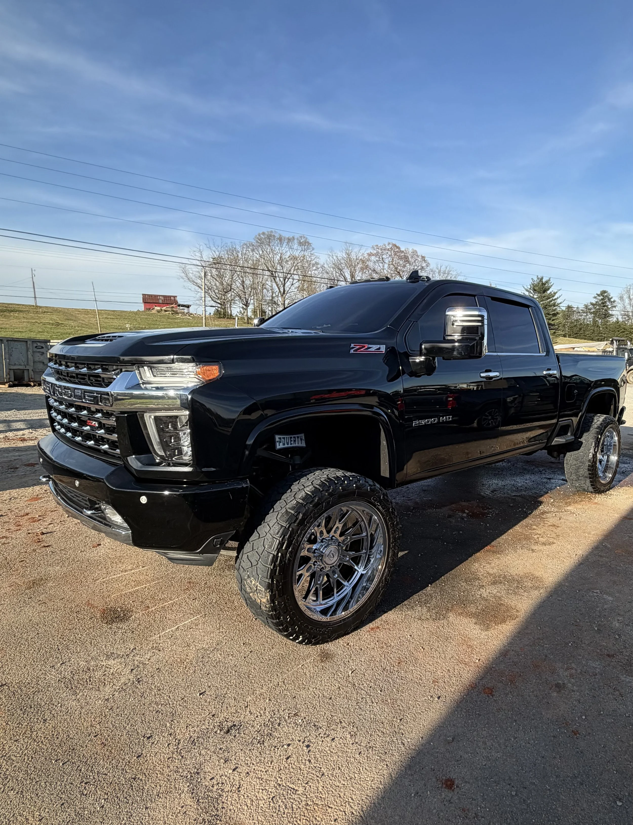 Front and side view of a black pickup truck parked on a gravel surface with trees, power lines, and a blue sky in the background.