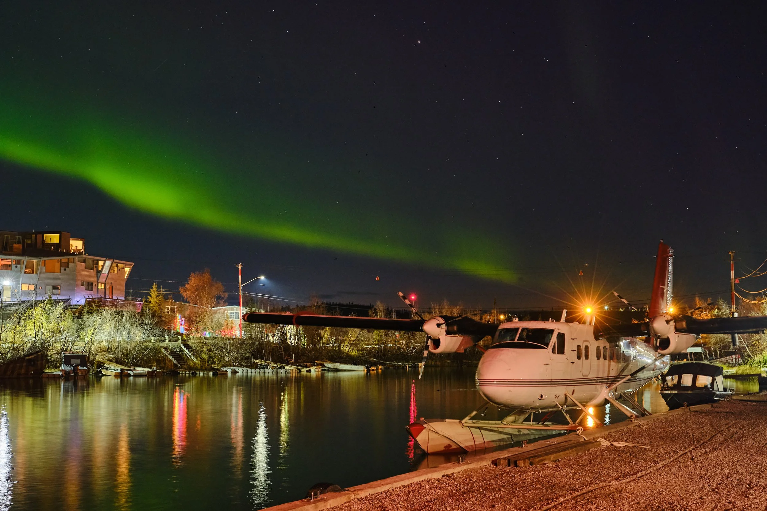Night scene of a marina with boats docked, a helicopter parked by the water, colorful northern lights in the sky, and buildings illuminated in the background.