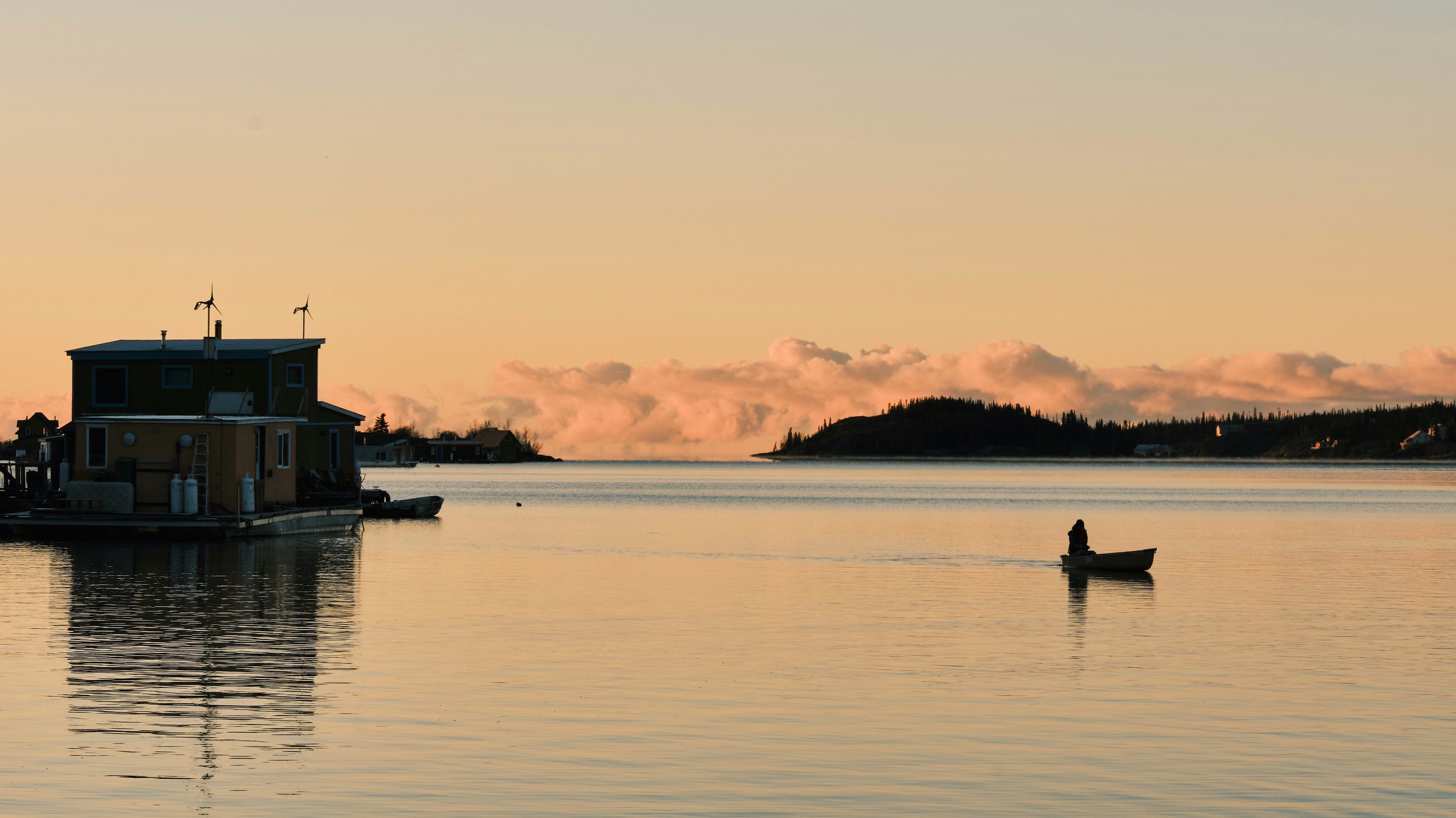 A calm body of water at sunset with a house on a boat and a person rowing a boat nearby, with hills and houses in the background.