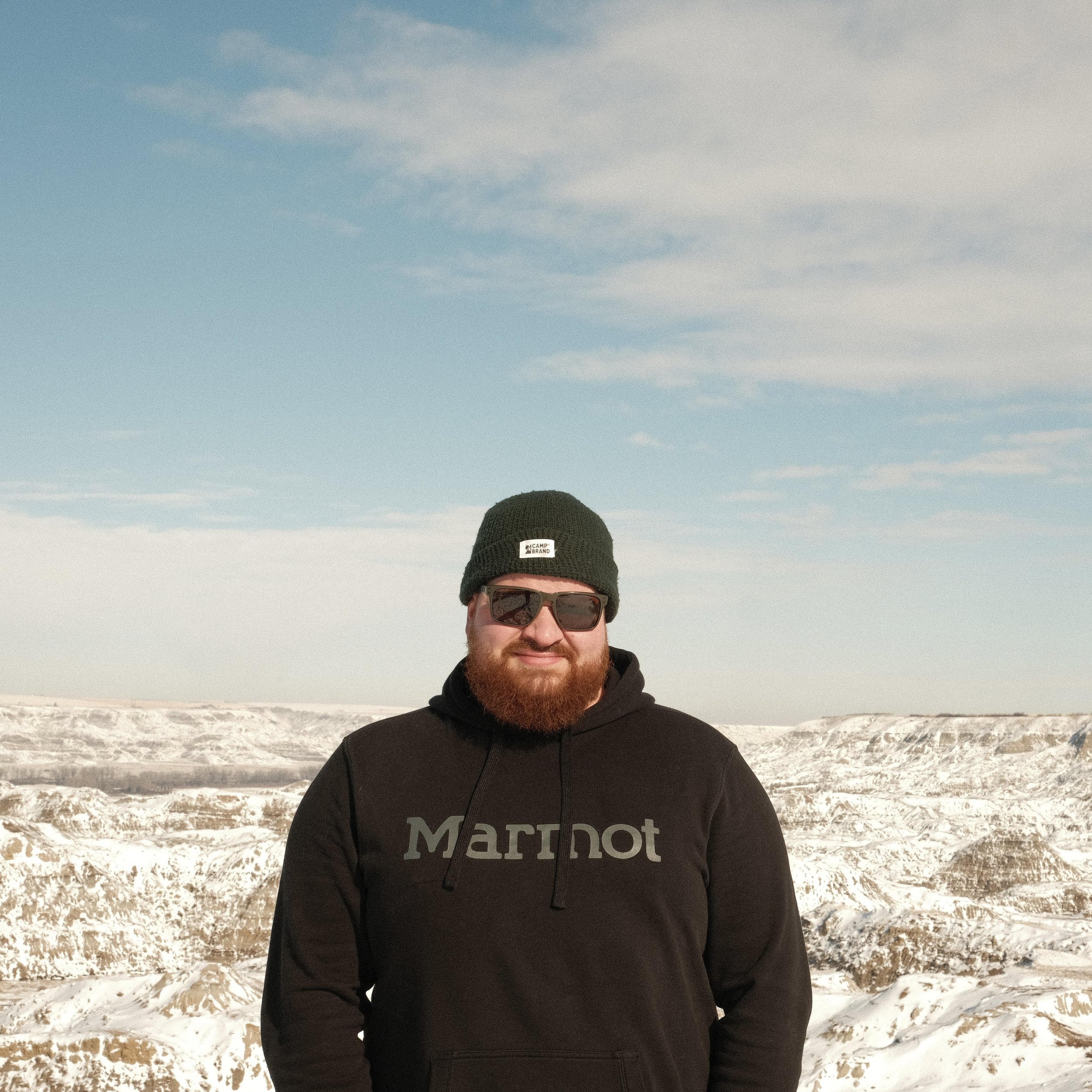 A man with a beard and sunglasses standing outdoors in a rocky, snow-covered landscape with mountain ridges in the background, wearing a black Marmot hoodie and a black beanie.