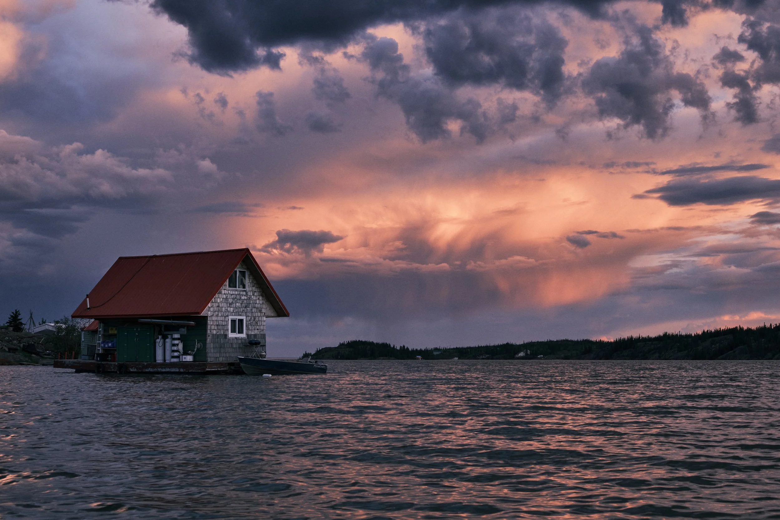 A small house on stilts by the water with a boat docked nearby, under a colorful, cloudy sunset sky.