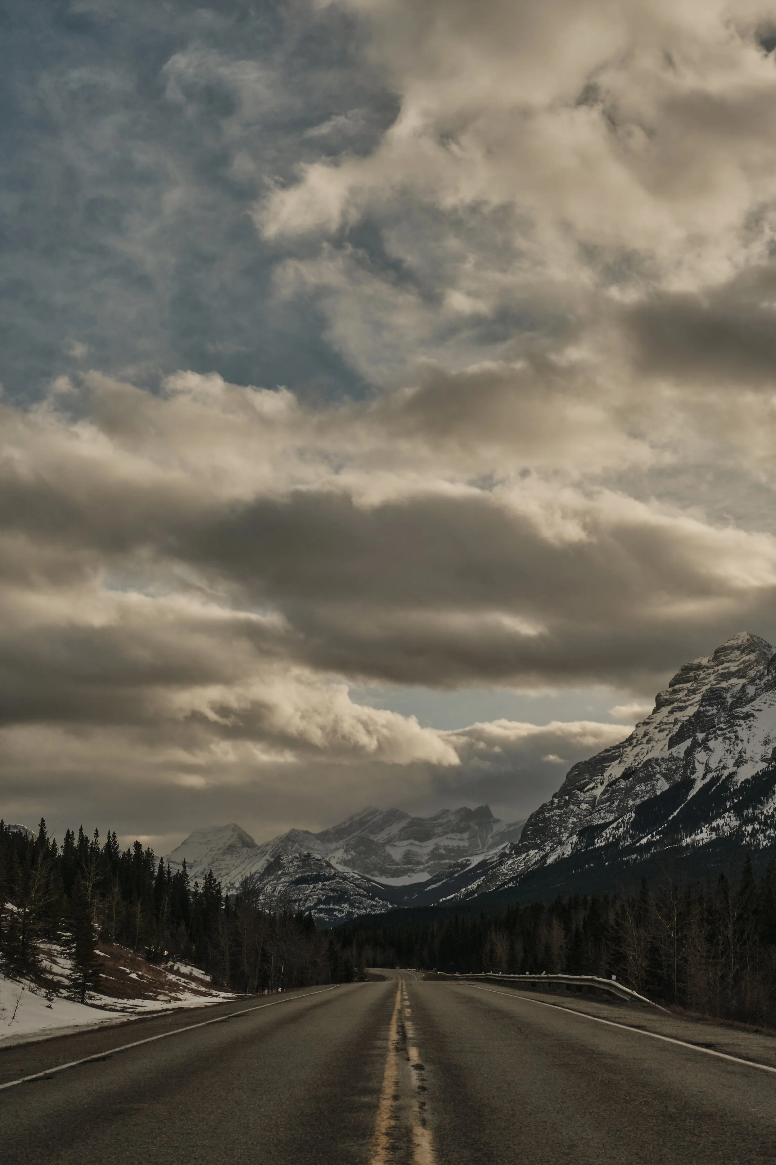 A mountain road with double yellow lines in the center, surrounded by snow-capped mountains, dense forests, and a cloudy sky.