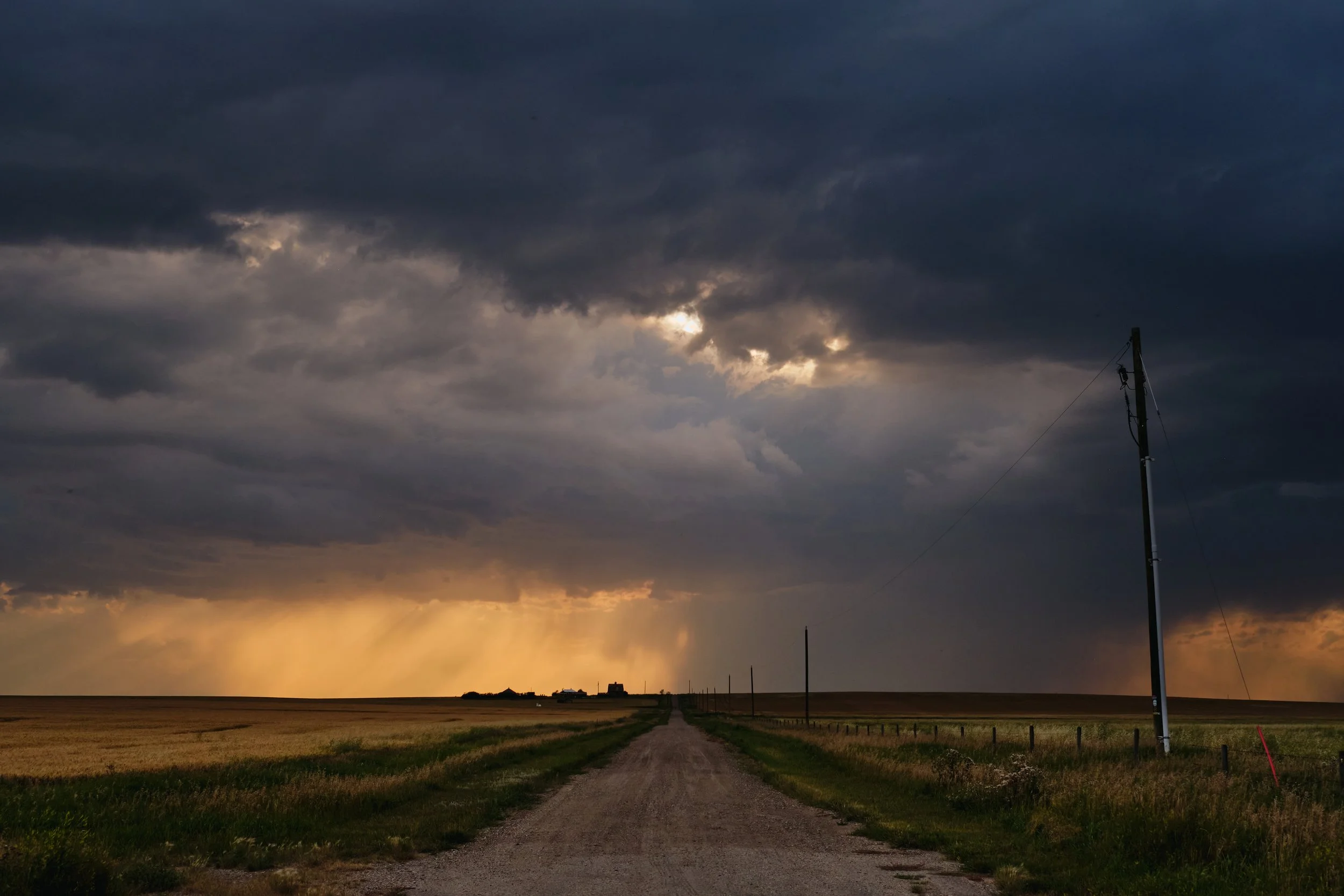 A rural landscape during sunset with dark storm clouds and a yellow glow on the horizon. A dirt road runs through the grassy fields, with utility poles and a small building far in the distance.