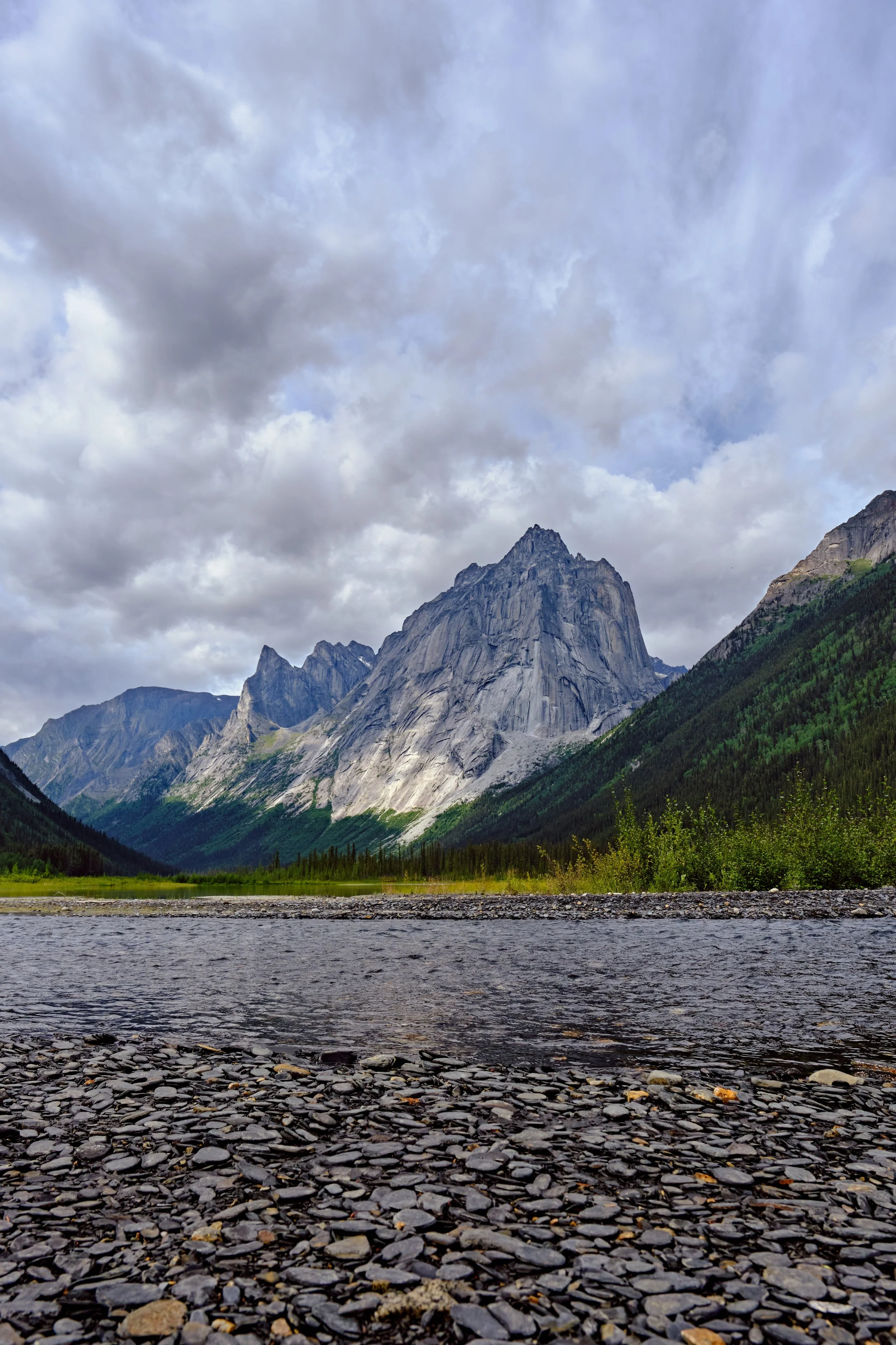 View of mountain range with cloudy sky, lush green forest, and a rocky riverbank in the foreground.