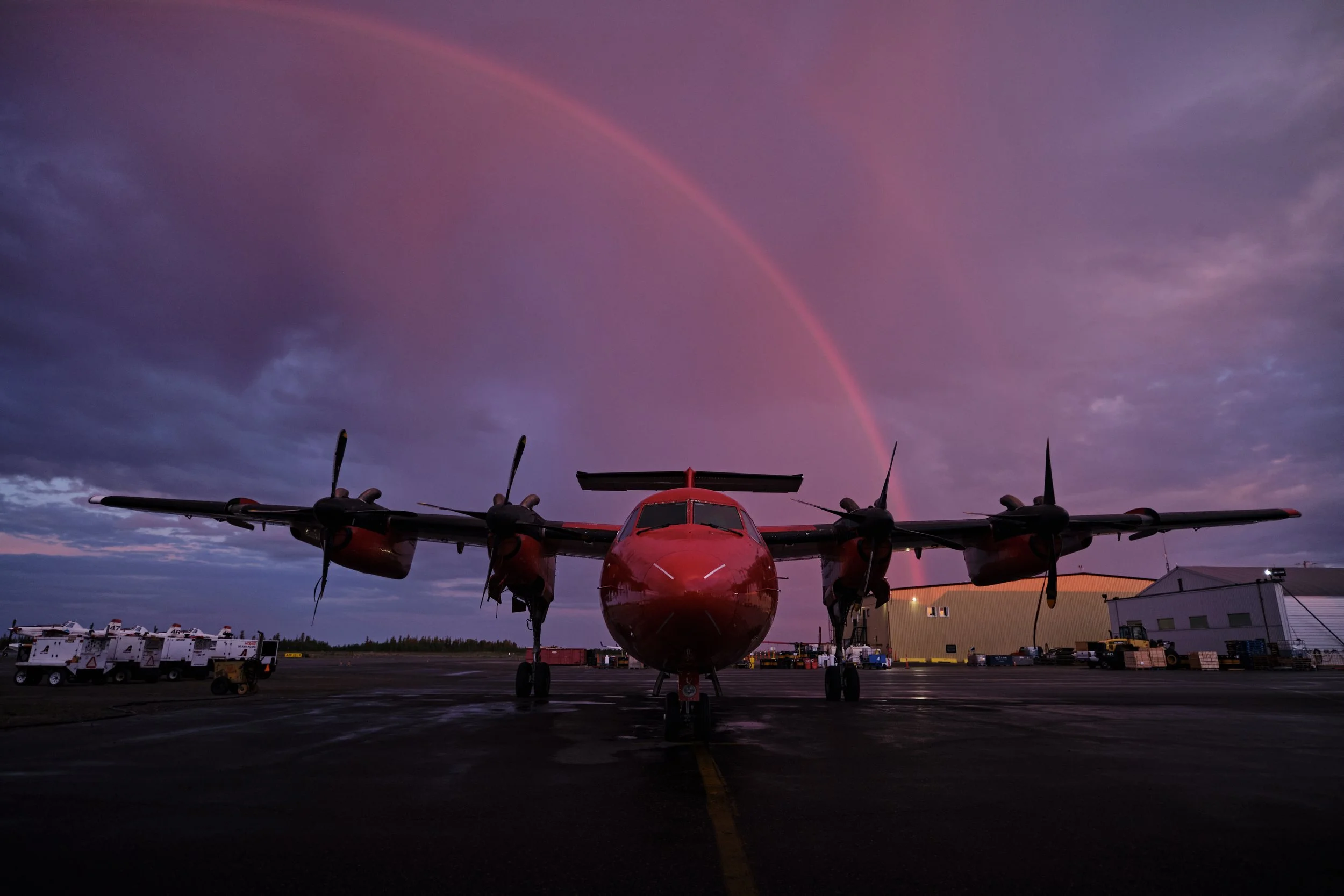 A red aircraft parked on the tarmac at dusk, with a rainbow in the purple sky above.
