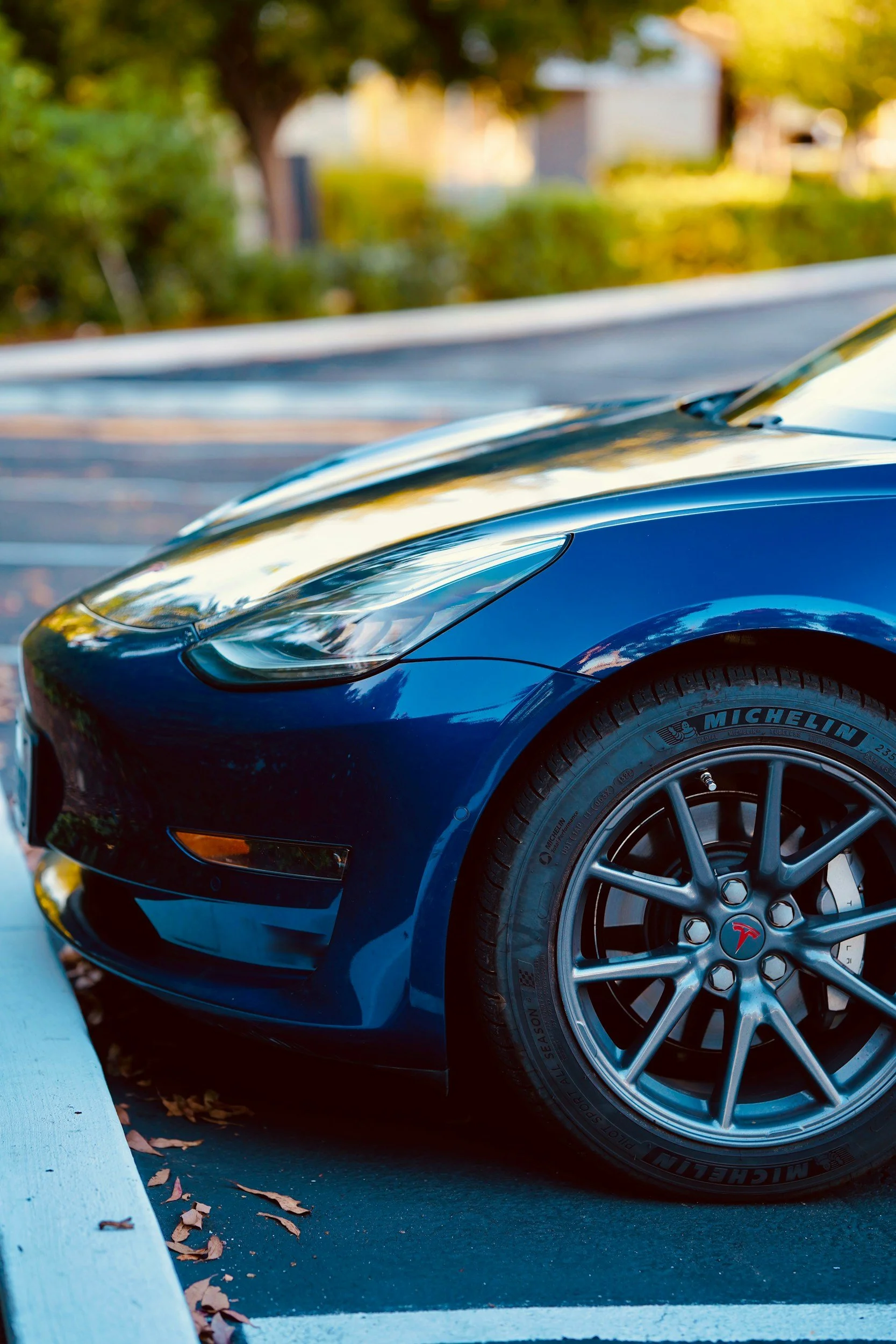 Close-up of a blue Tesla Model S parked in a parking lot with a curb and fallen leaves nearby, with trees and buildings in the background.