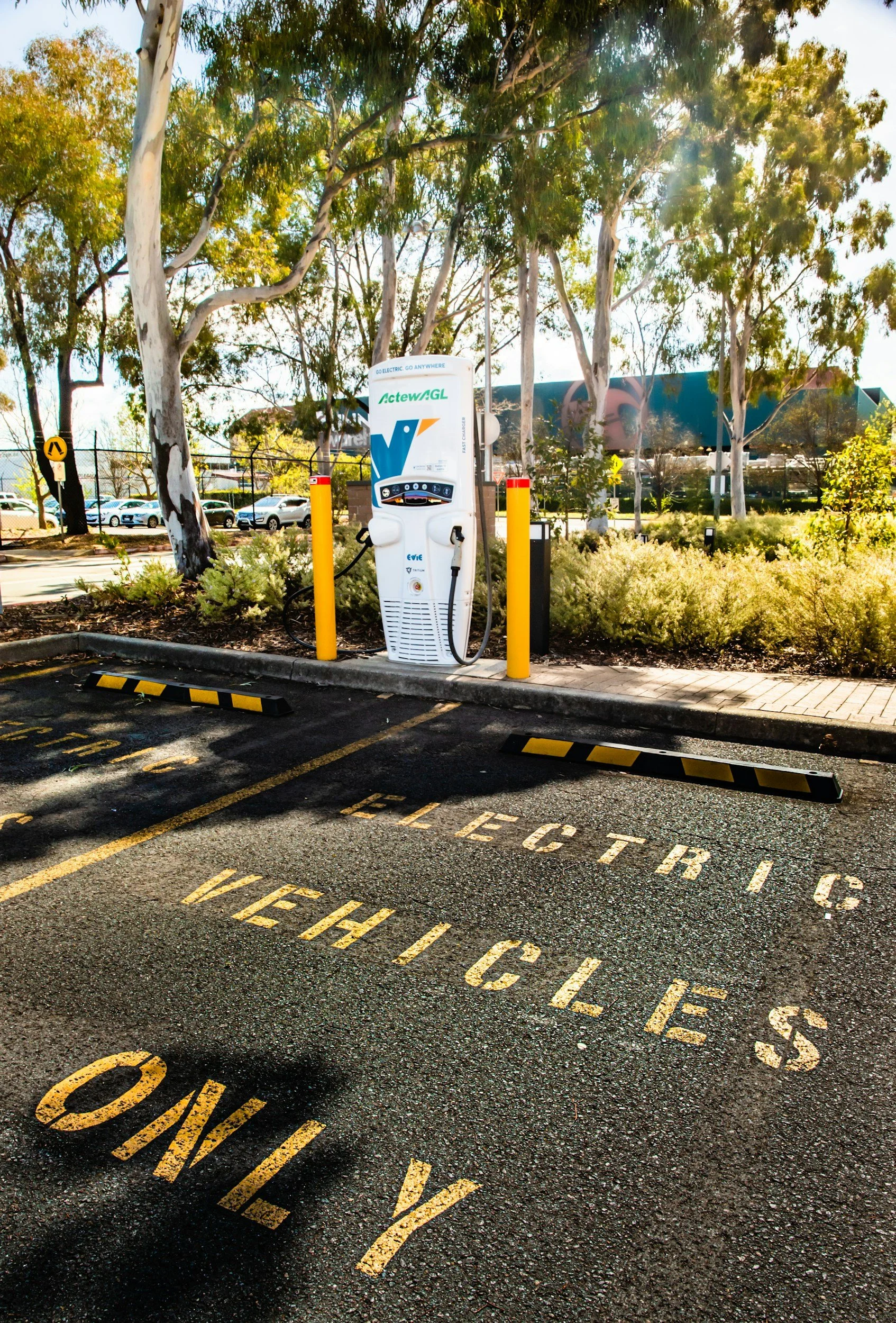 Electric vehicle charging station in a parking lot, surrounded by trees and plants, with parking space markings on the ground reading 'ELECTRIC VEHICLES ONLY.'