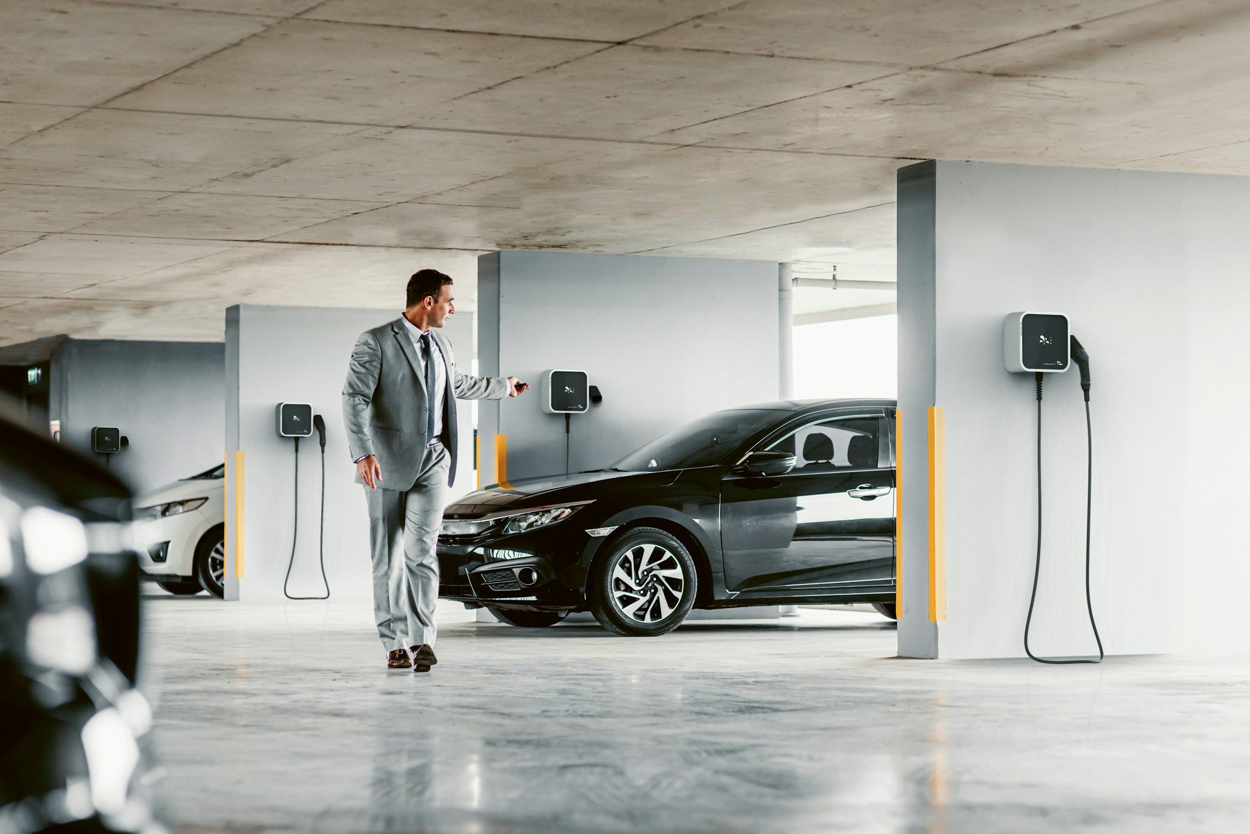 A man in a gray suit is charging a black electric car in an indoor parking garage.