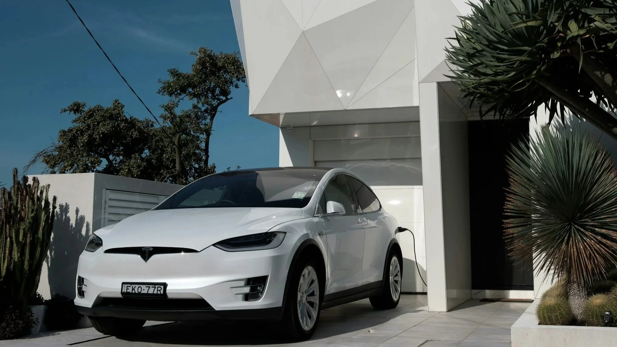A white Tesla Model Y electric car charging outside a modern white house with greenery and trees against a blue sky.