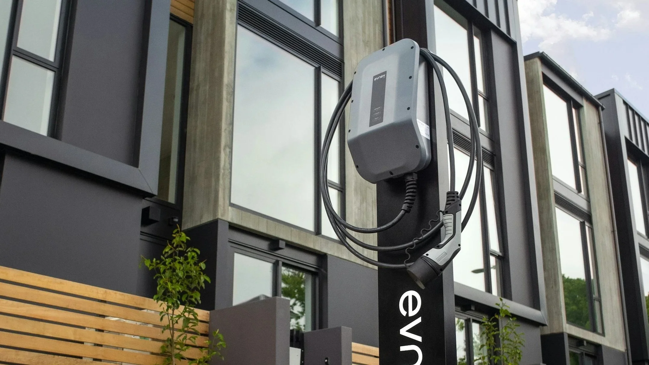 Electric vehicle charging station mounted on a black pole in front of a modern building with large glass windows and wood paneling, with some greenery at the bottom.