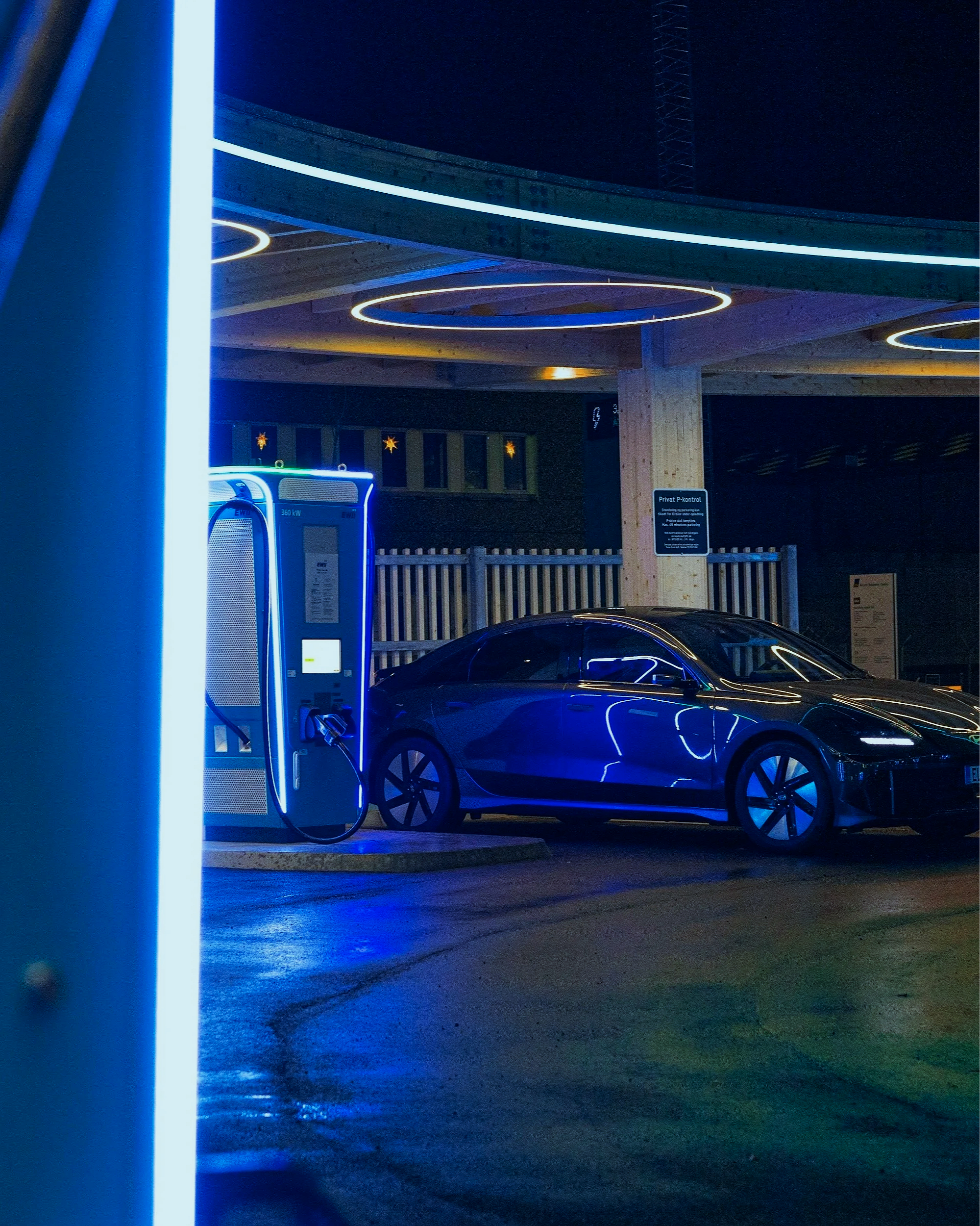 A sleek electric car is parked next to an electric charging station at night, illuminated by blue neon lights.