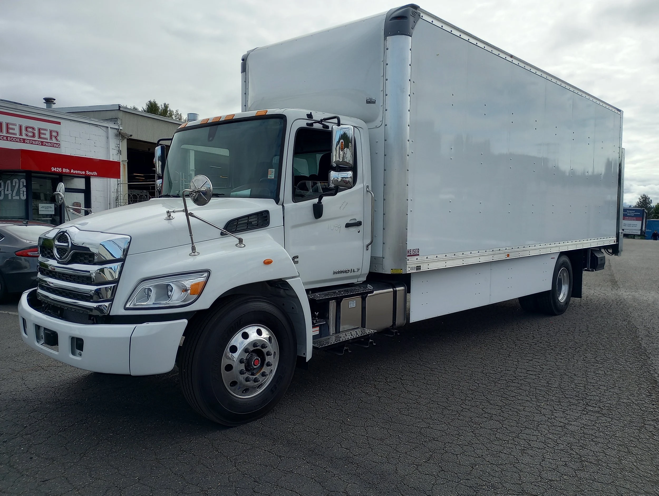 A custom installed white box truck parked on a paved lot, with the Heiser Body building and cars in the background.