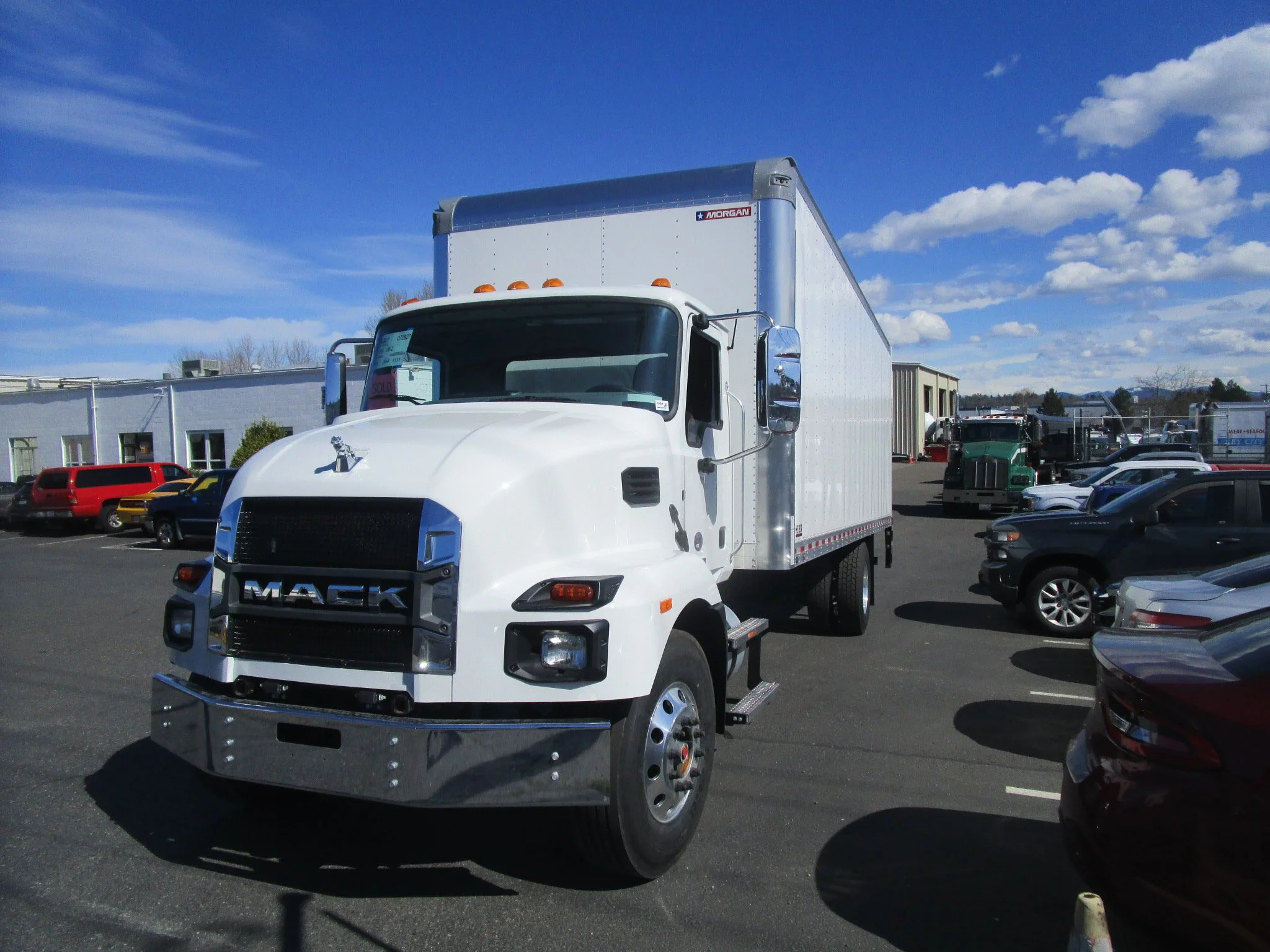 White Mack box truck parked in a lot with other vehicles and a blue sky with scattered clouds.