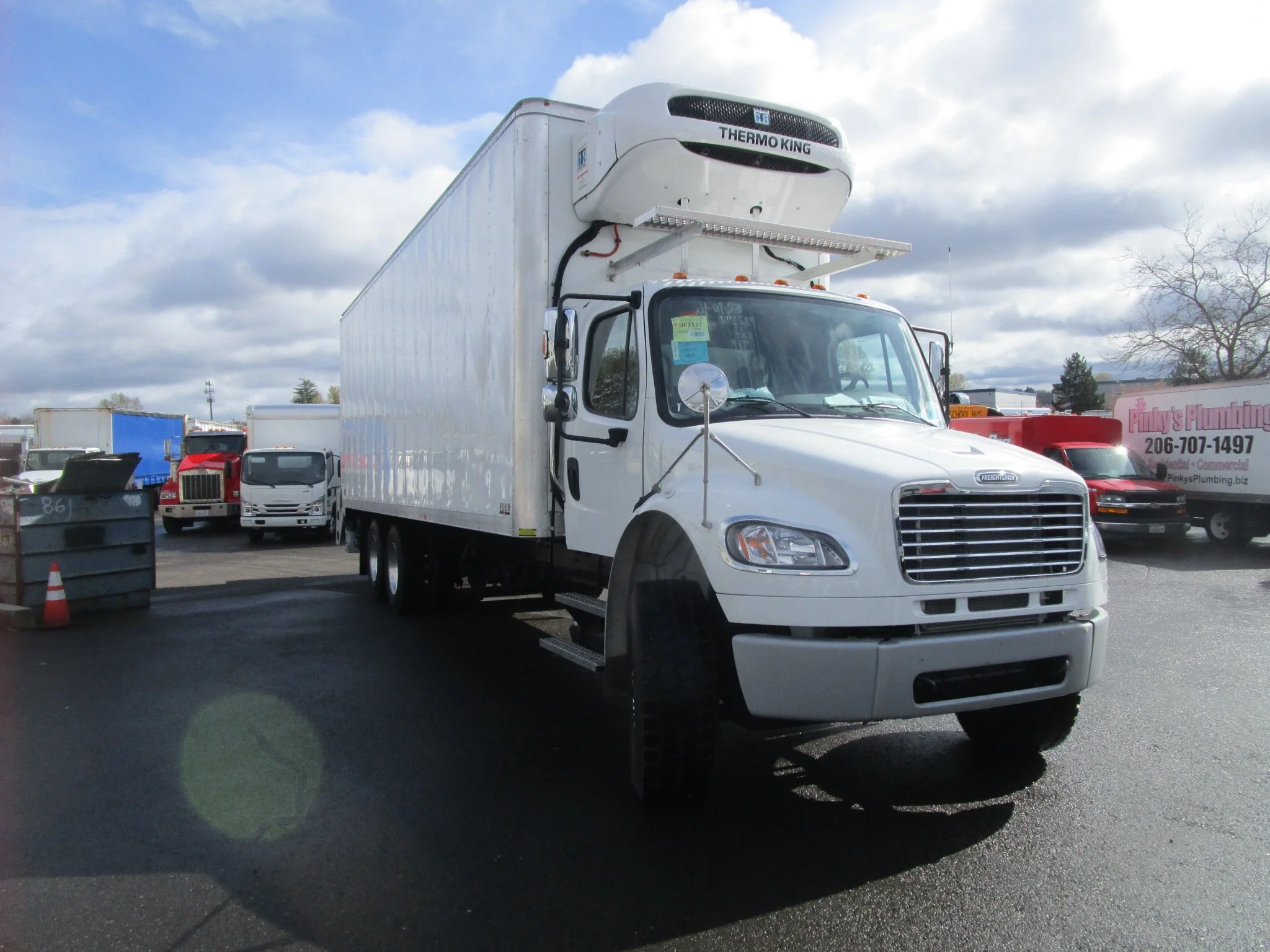 White delivery truck with a custom installed refrigerated box and Thermo King unit on top parked on a lot with other trucks and vehicles under cloudy skies.