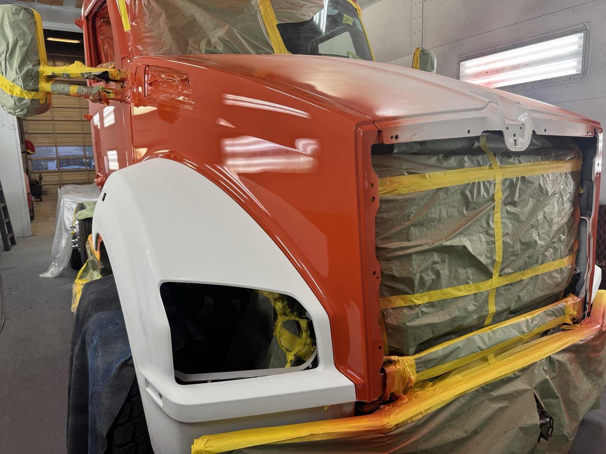 A vehicle undergoing paint preparation inside the Heiser Body workshop, with parts masked off and covered in brown paper and yellow tape, showing a red front hood and a white front fender.