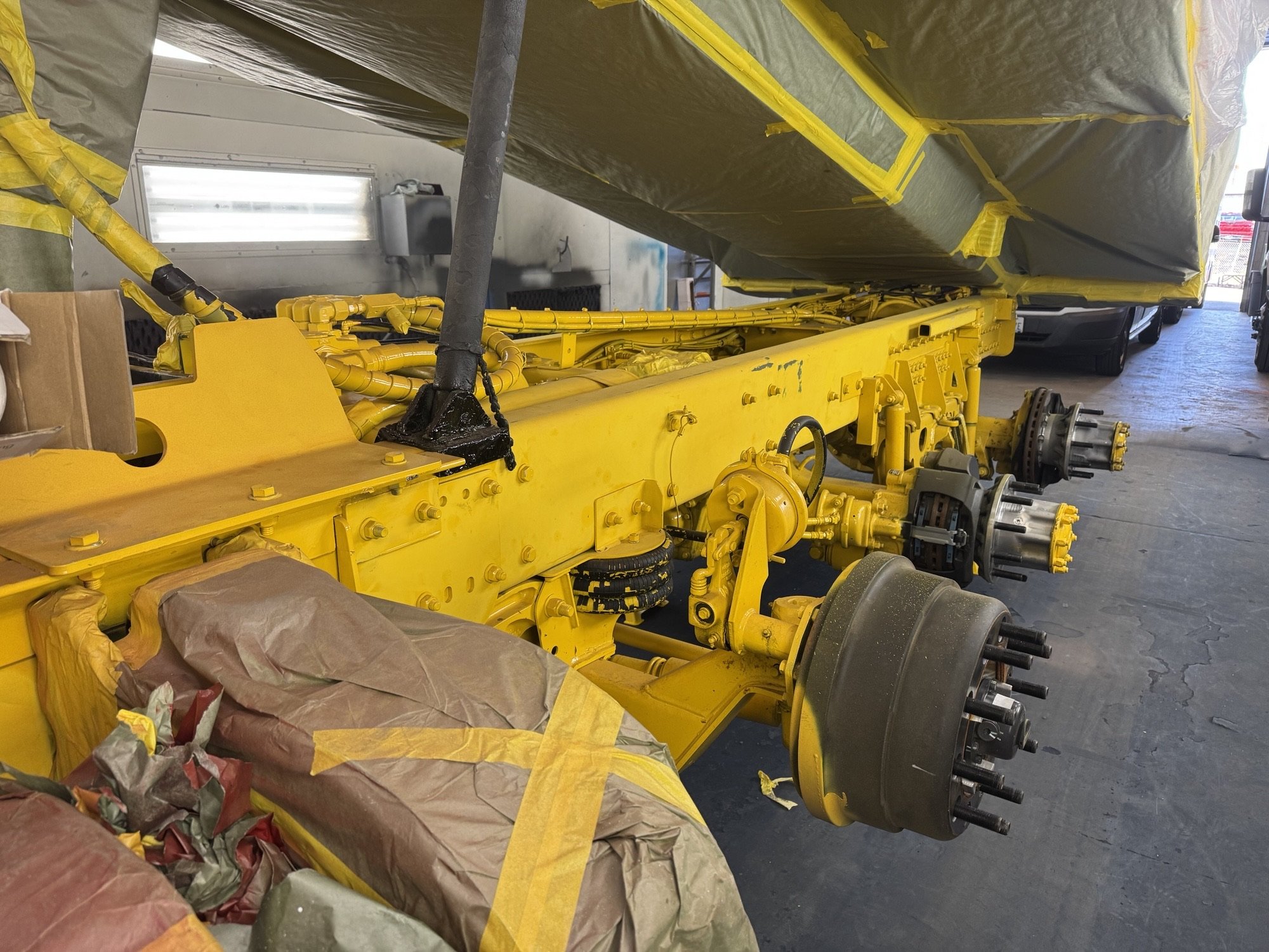 A yellow-painted vehicle frame without wheels, with visible hydraulic and mechanical components, is in a workshop or garage. The frame is elevated and partially covered with plastic and paper masking materials, suggesting it is undergoing maintenance or painting.