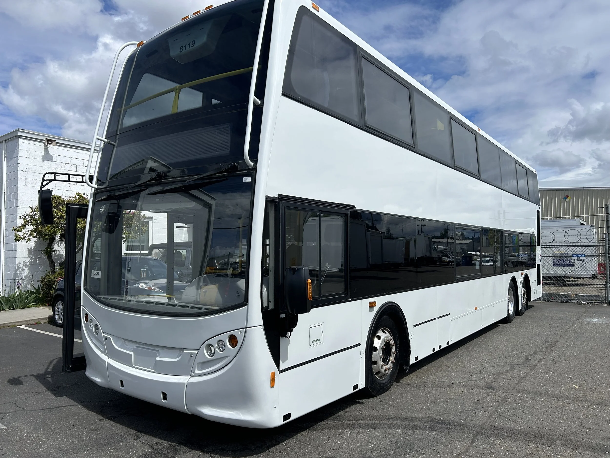 Freshly painted white double-decker bus parked in a lot under a partly cloudy sky.
