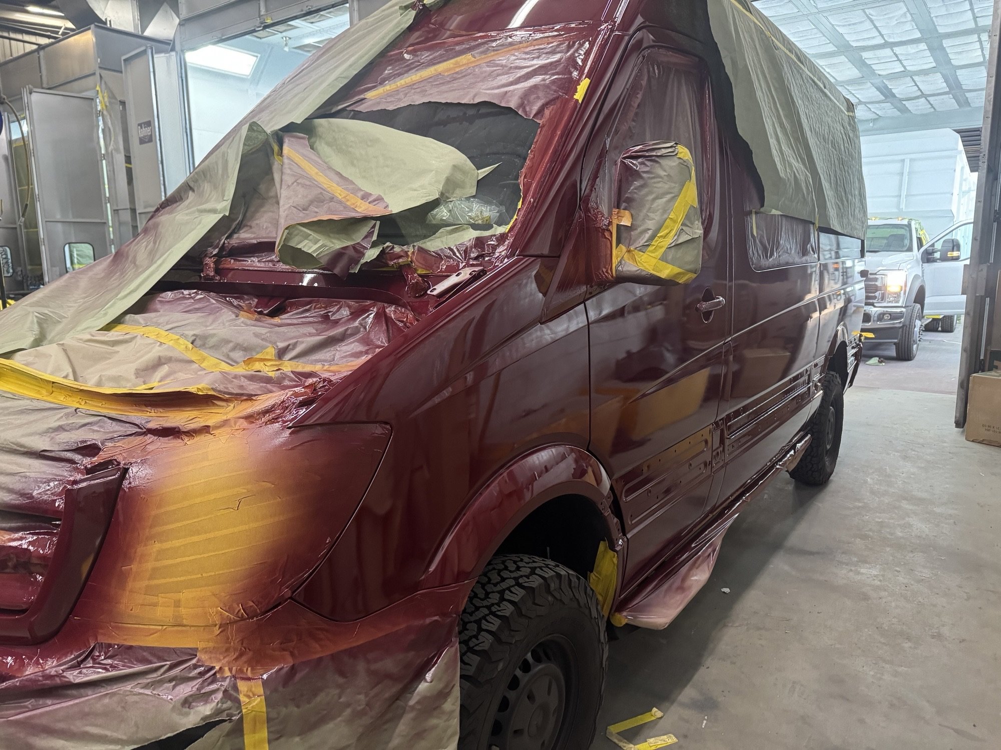 A maroon van being prepped for painting by Heiser Body with masking tape and plastic covers. Another vehicle is visible in the background.