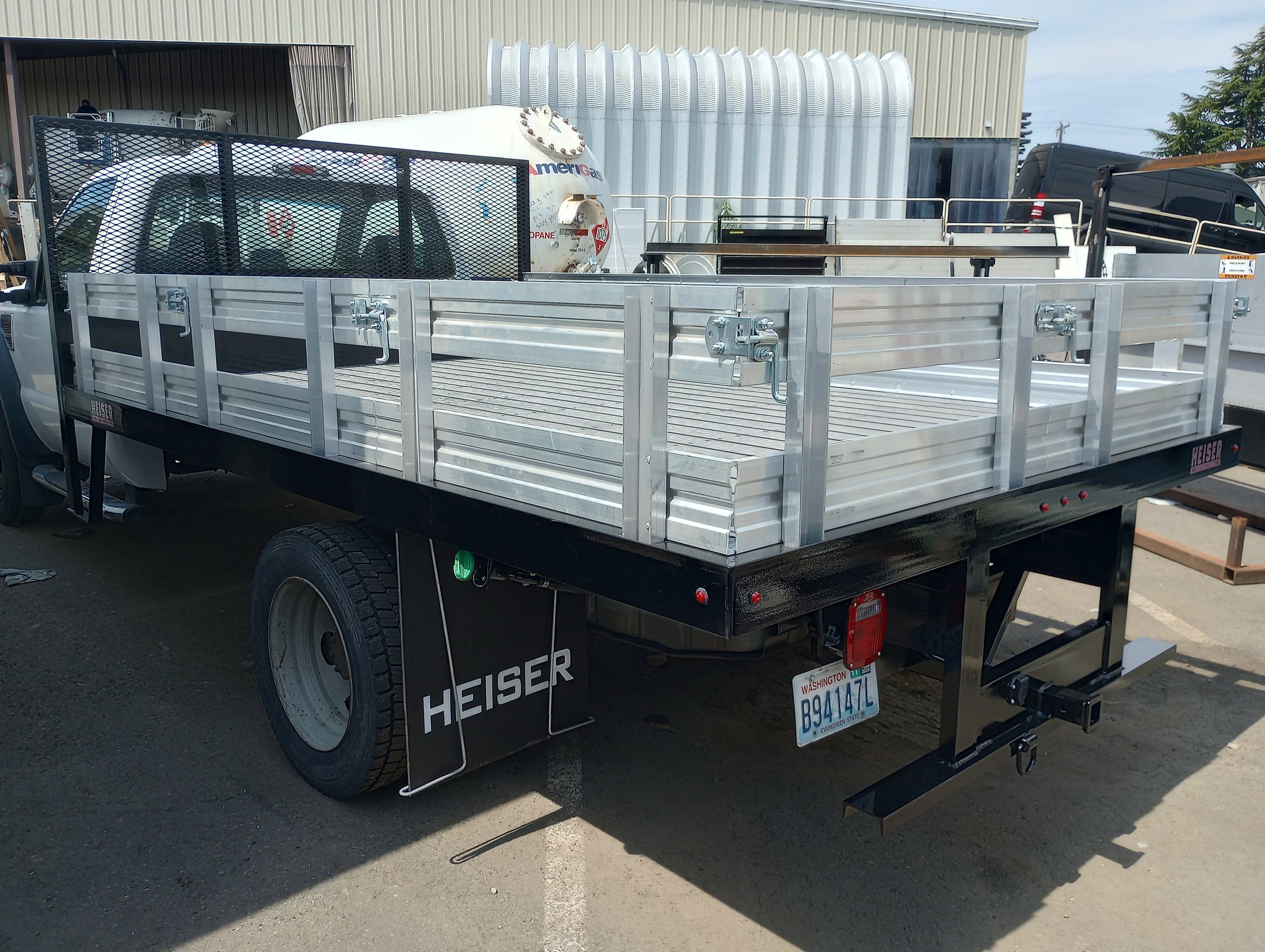 A custom built flatbed utility trailer with a metal frame, sides, and a black chassis, attached to a pickup truck, parked outdoor Heiser Body. 