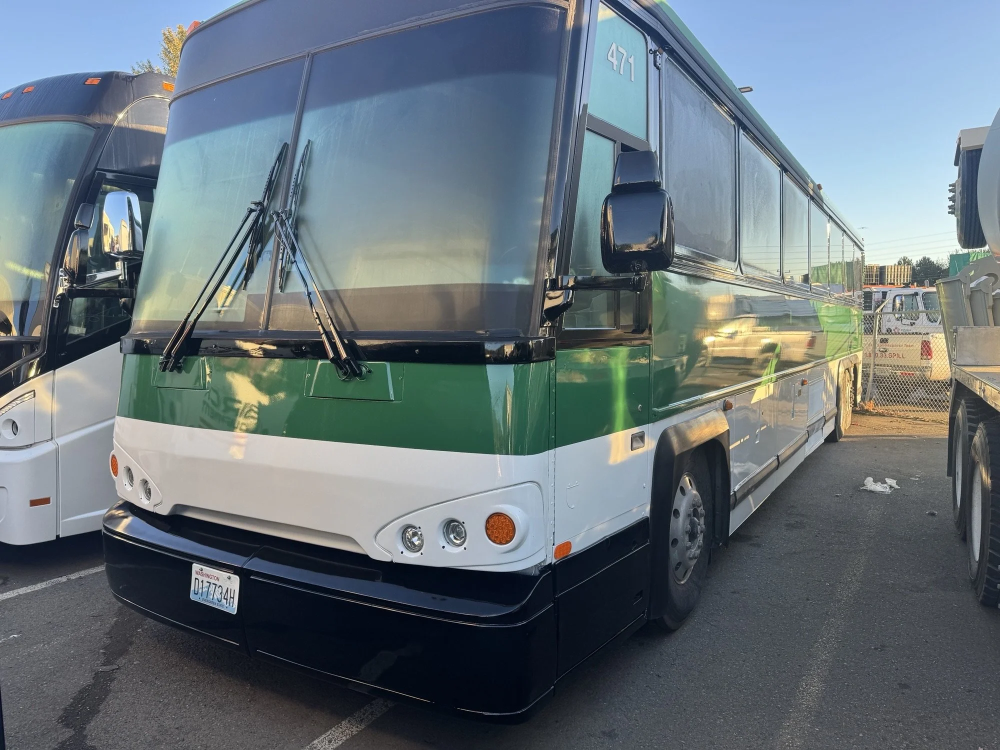 A freshly painted large bus with a green and white color scheme, parked in an outdoor lot near other buses and vehicles, with a chain-link fence in the background.
