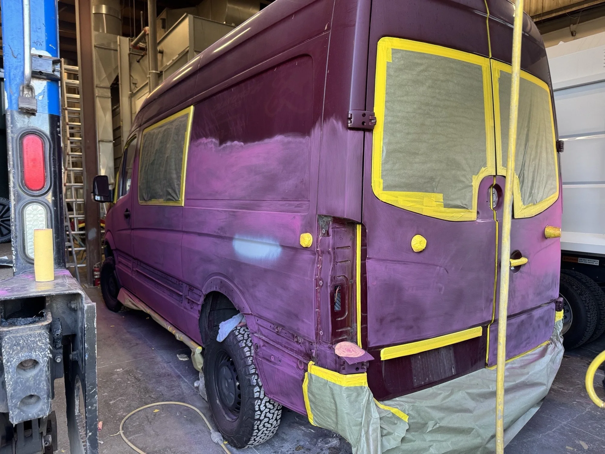 A van being painted purple by Heiser Body inside their workshop with masking tape on windows and edges, and covered tires.