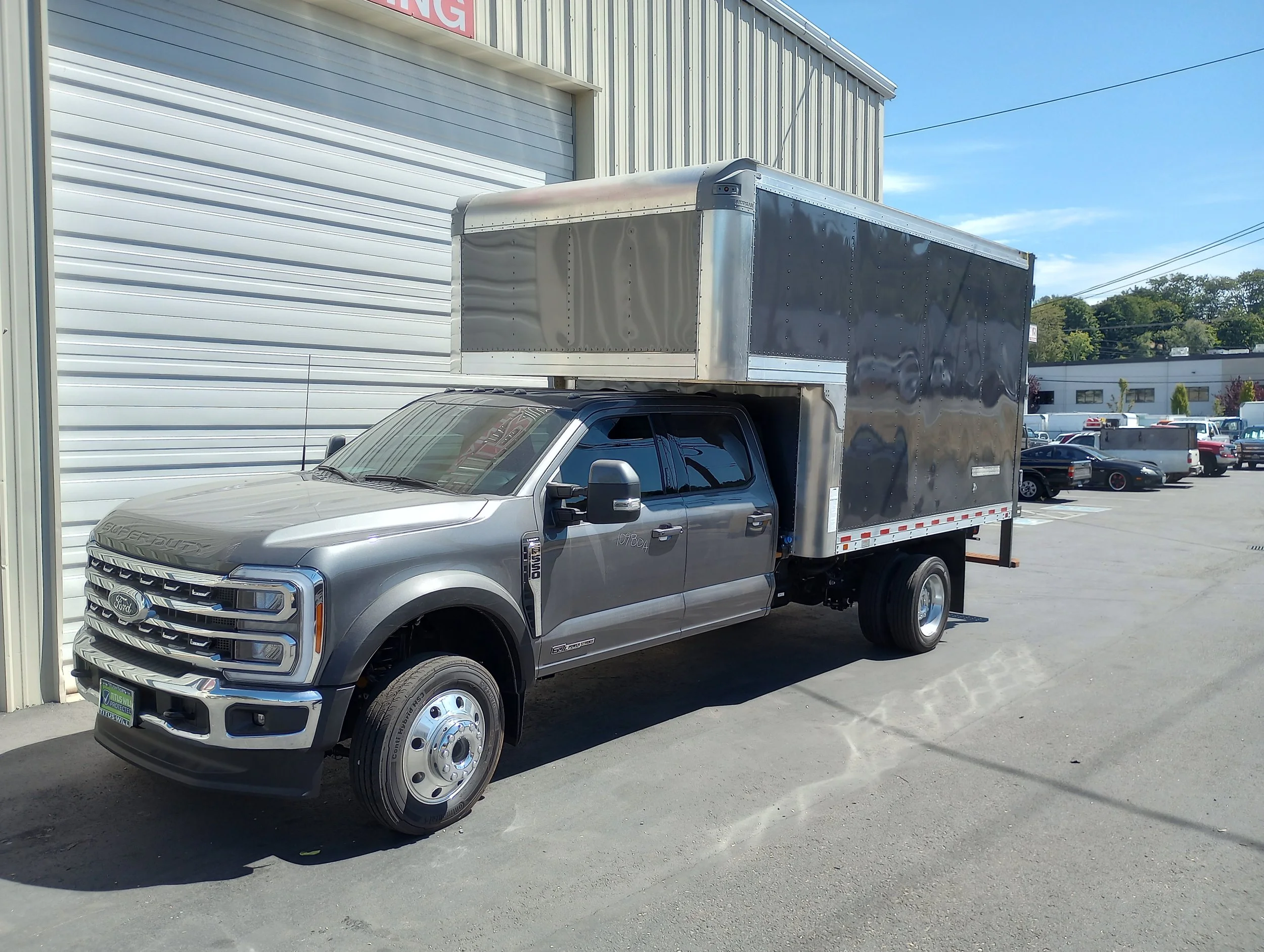 Silver Ford truck with custom installed black cargo box parked outside the Heiser Body building on a sunny day. In the background, there is a parking lot with several other vehicles.