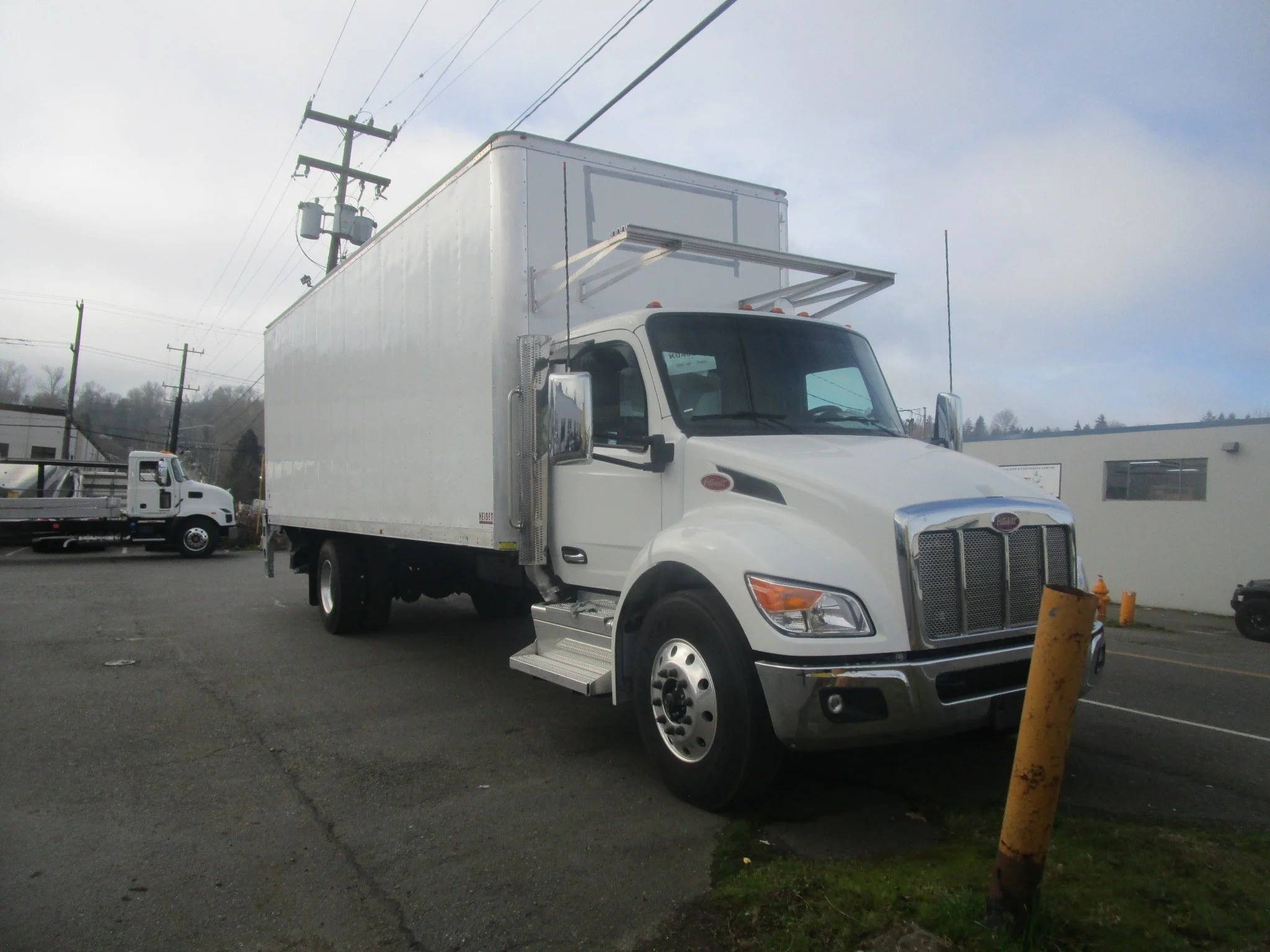A custom installed white box truck parked next to a yellow pole on a paved lot with utility poles and the Heiser Body building in the background.