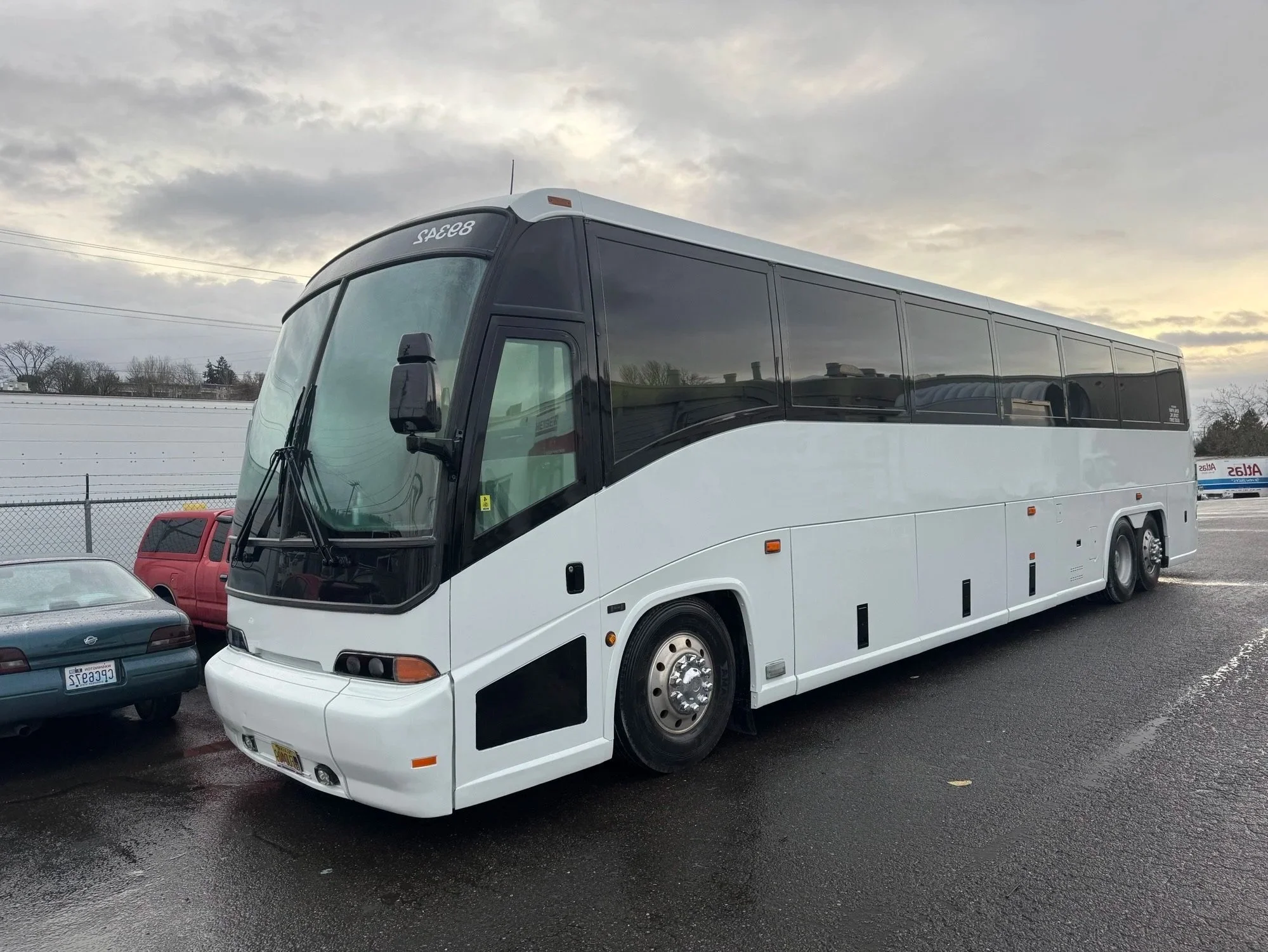 White and black coach bus parked in the Heiser Body parking lot with other vehicles, cloudy sky in the background.
